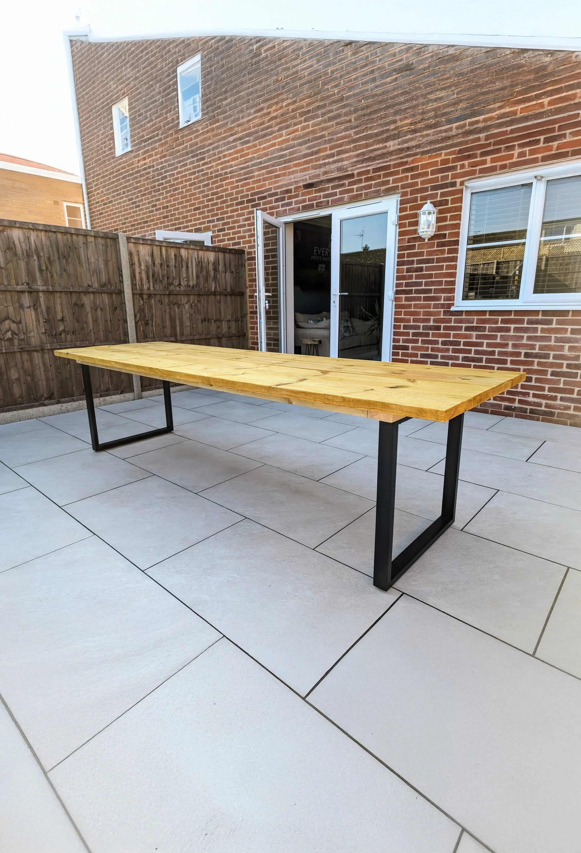 A wooden outdoor table with black metal legs on a tiled patio with a brick house in the background, a sliding glass door, and a wooden fence.