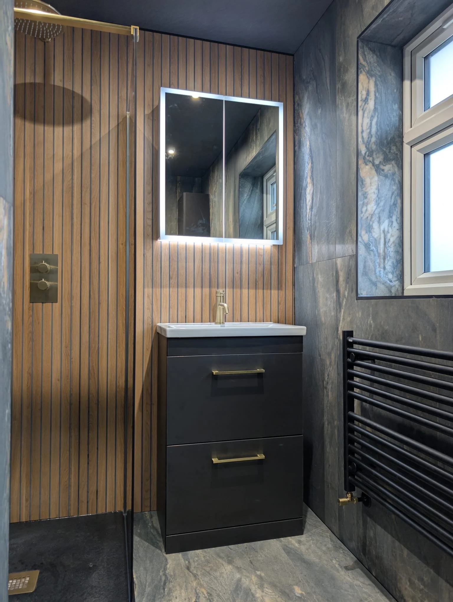 Modern bathroom with wooden and gray stone walls, a black vanity with brass handles, a mirror with backlit LED lighting, small window, and black heated towel radiator.