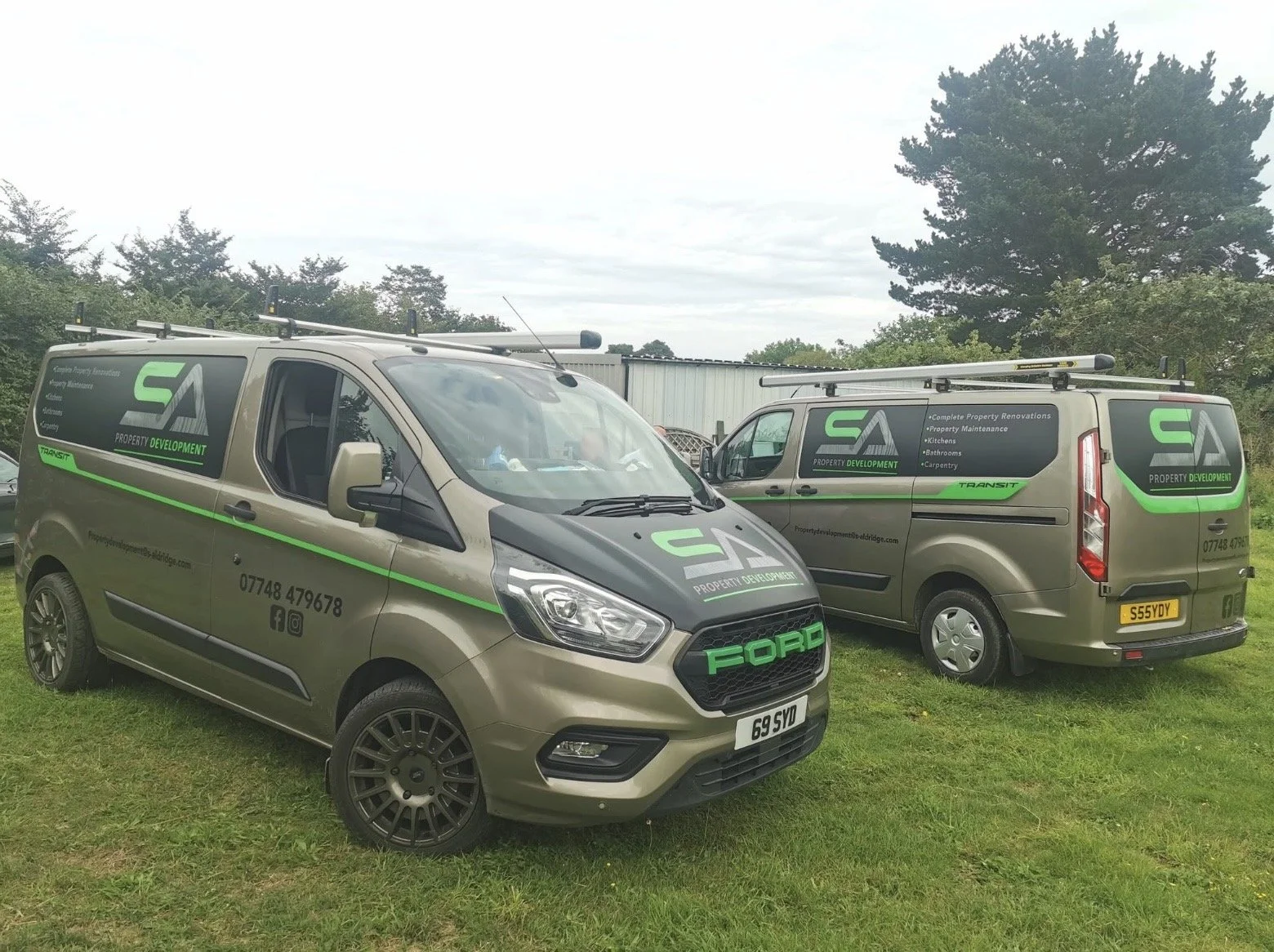 Two beige company vans with green and black advertising decals parked on grass in an outdoor setting. The vans display the logo and contact information for Property Development, showing services like renovations, maintenance, kitchens, bathrooms, and carpentry. One vehicle is a Ford Transit, and the other is a larger van, both equipped with roof racks.