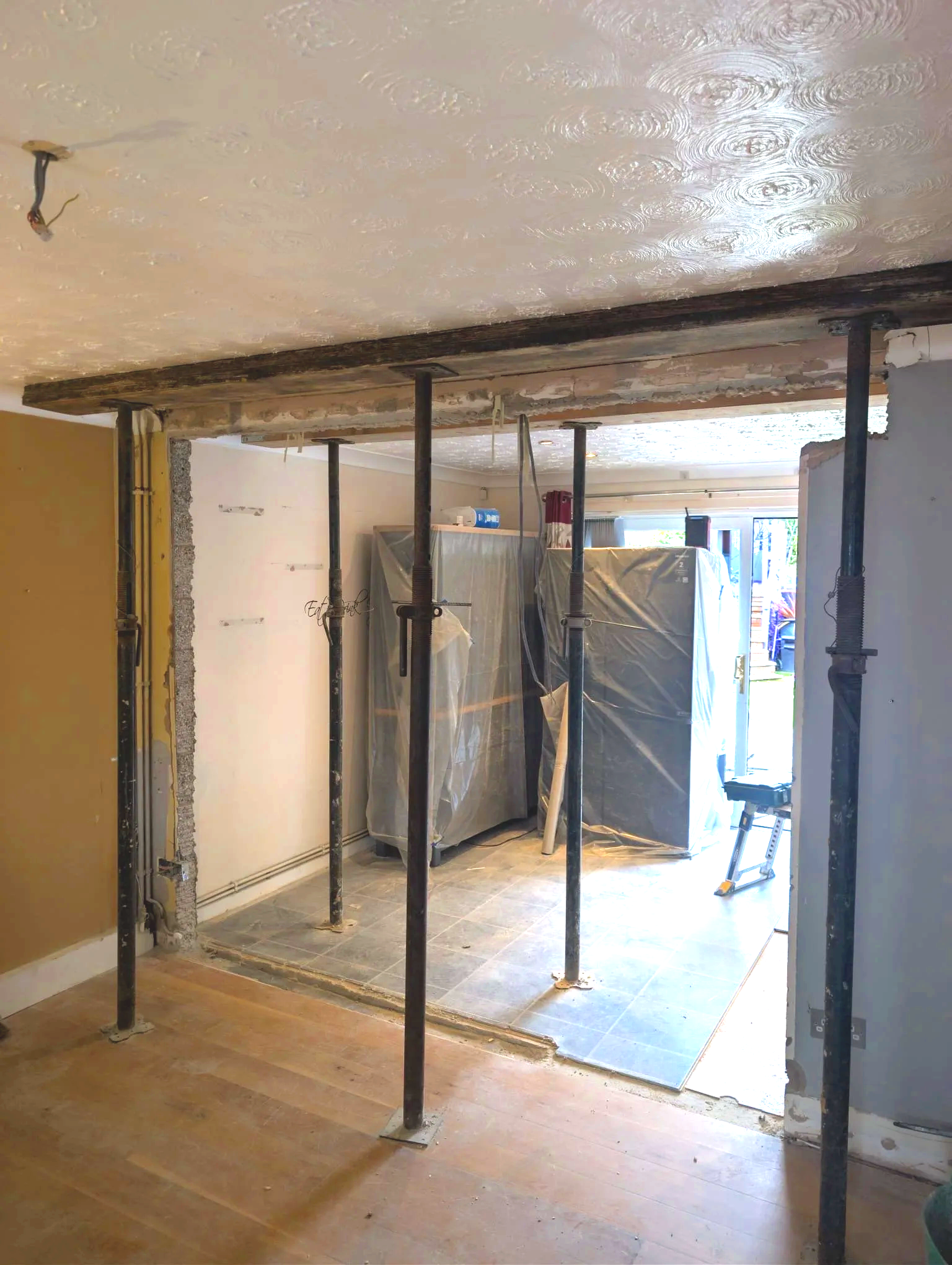 Room under renovation with support poles, partially demolished wall, and a construction area with plastic-covered appliances in the background.