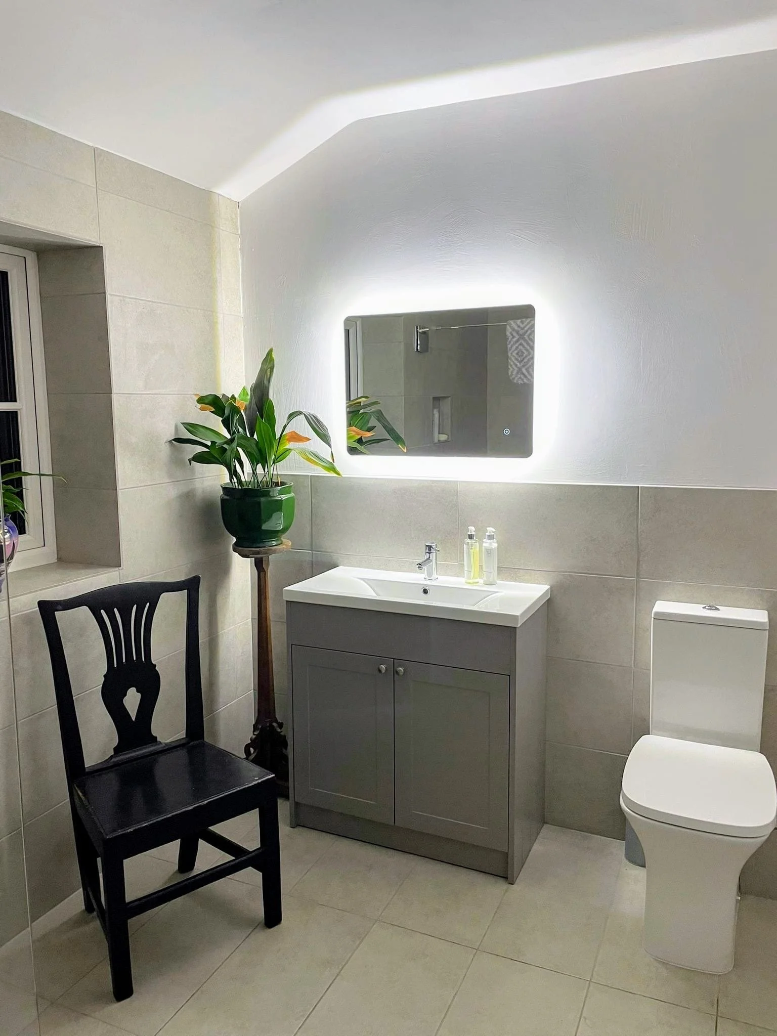 A modern bathroom with a white toilet, a small gray vanity with a white sink, and a rectangular mirror with backlit lighting. There is a black wooden chair and a tall plant in a green pot near a window. The bathroom has light-colored tiled walls and floor.