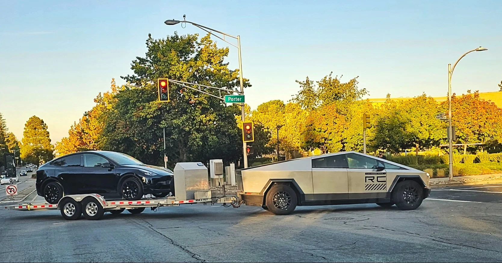 A black Tesla Model 3 being towed on a trailer attached to a futuristic, Tesla Cybertruck in an intersection with traffic lights, street signs, and trees during the daytime.