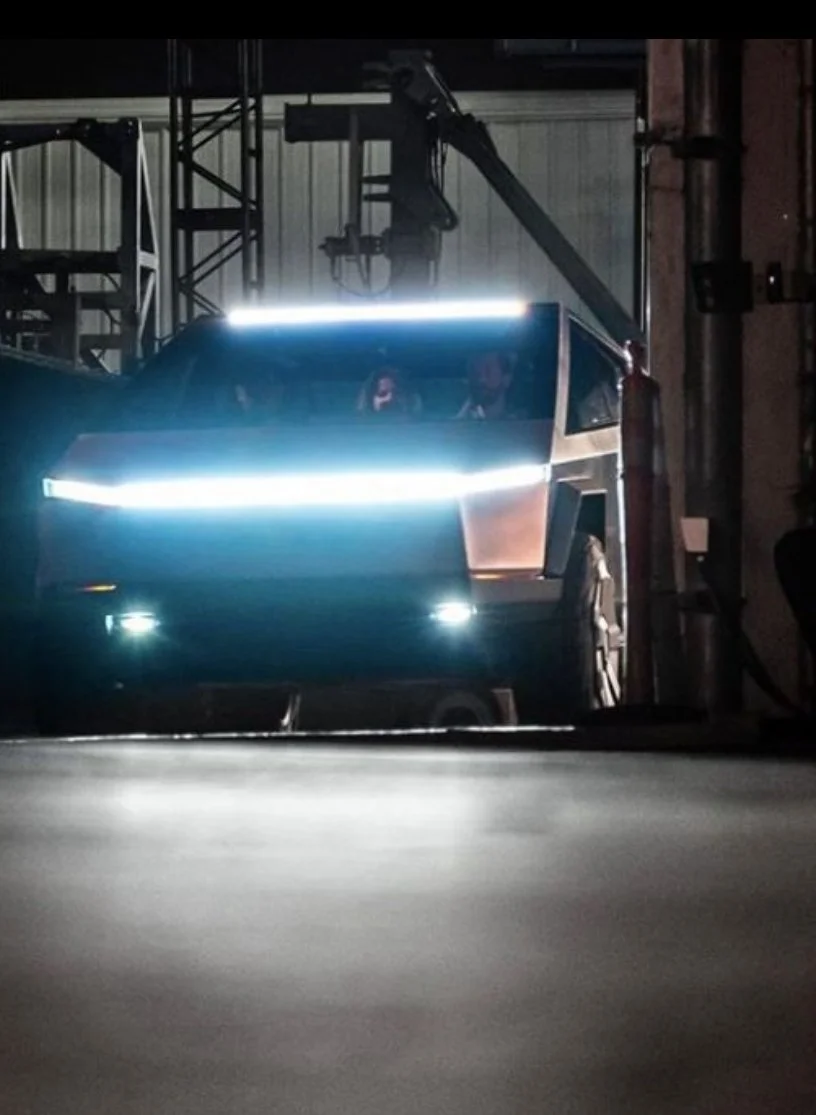 A futuristic, sci-fi style vehicle with bright LED lights on the front, inside an industrial setting with metal beams and a crane overhead, with two people visible inside.
