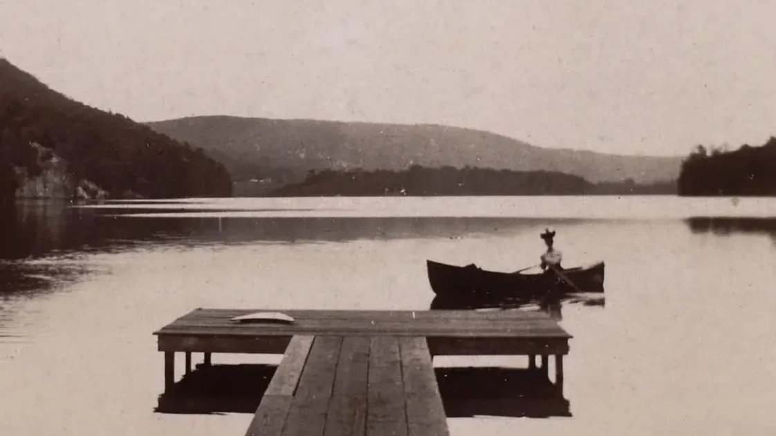A wooden dock extends into a calm lake with a person rowing a small boat nearby. Hills surround the lake in the background.