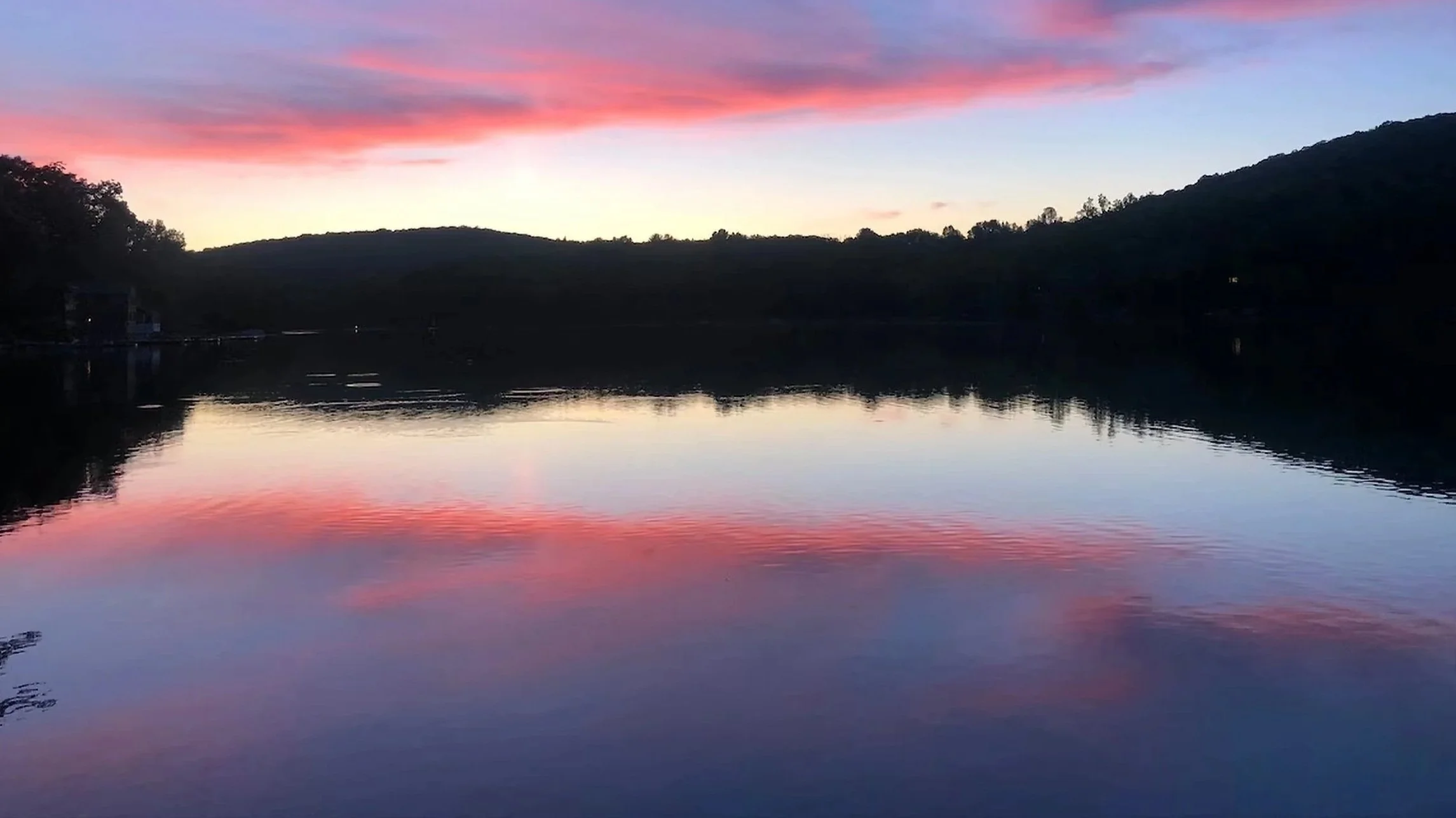 A peaceful lake at sunset with colorful pink and blue clouds mirrored on calm water, surrounded by dark hills.