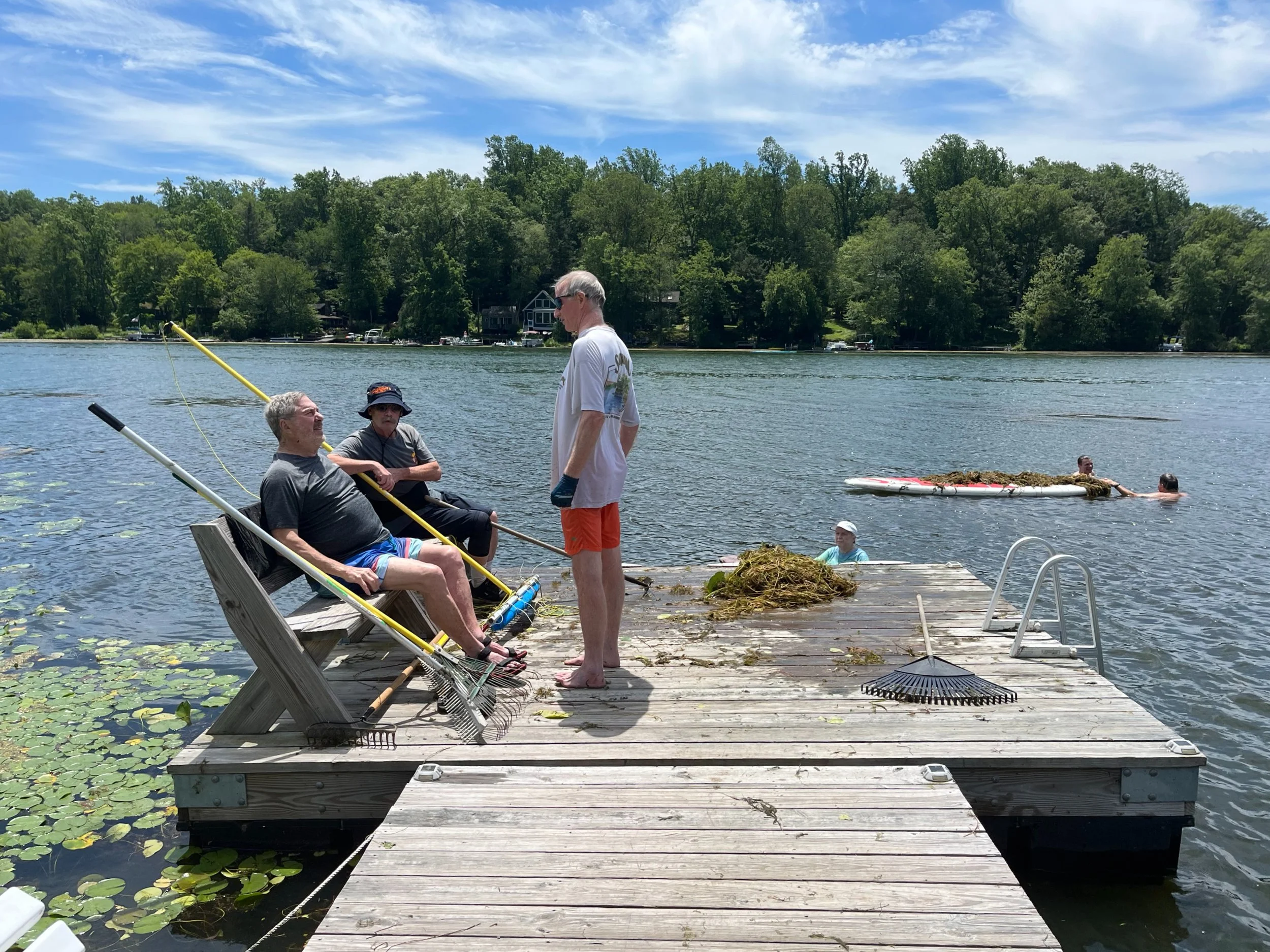 Group of people on a wooden dock by a lake, with some sitting and others in the water, surrounded by trees and blue sky.