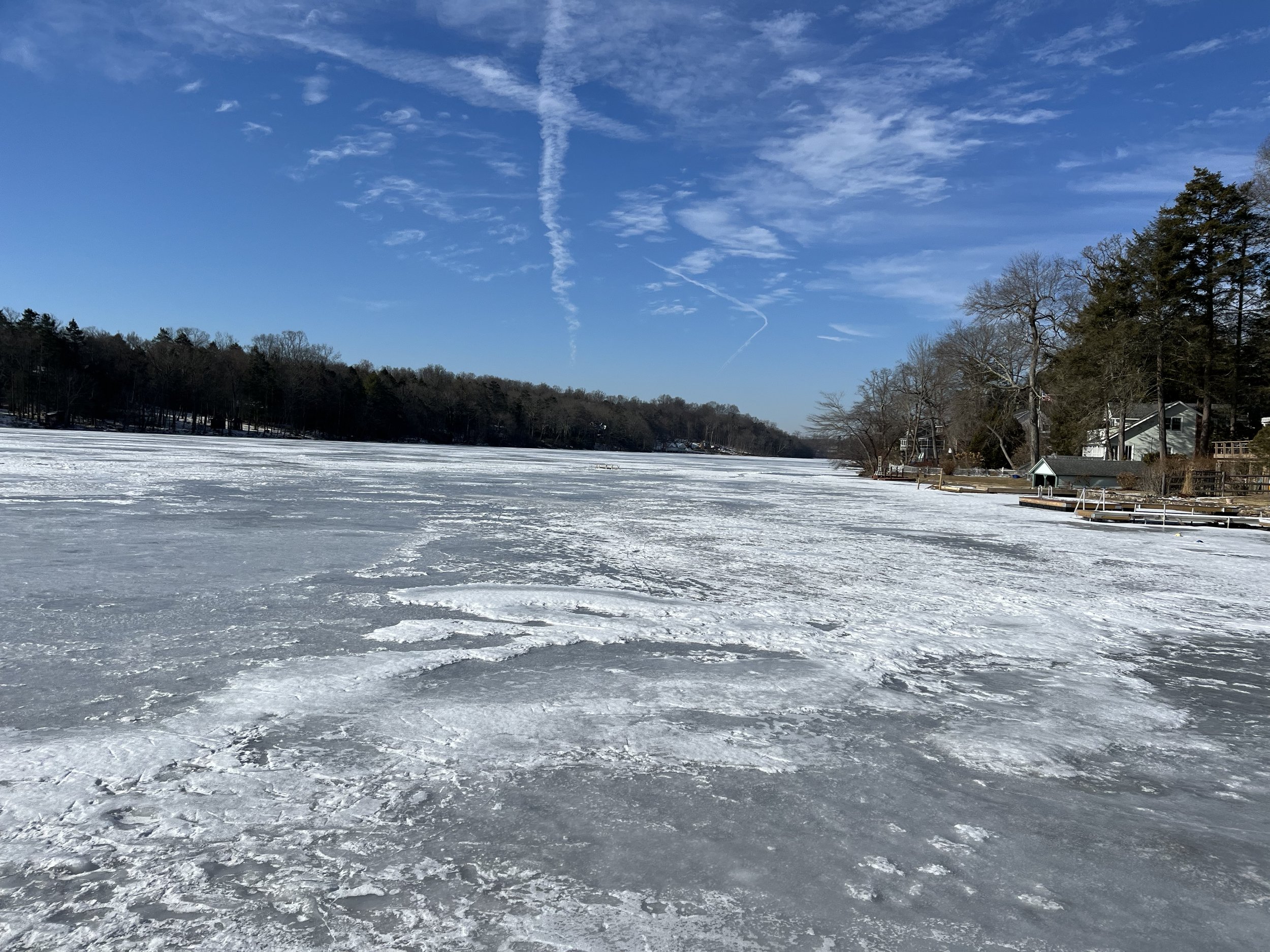 Frozen lake with ice and snow, surrounded by trees and houses, under a blue sky with wispy clouds.