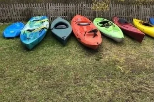 Six colorful kayaks resting on grass in front of a wooden fence.