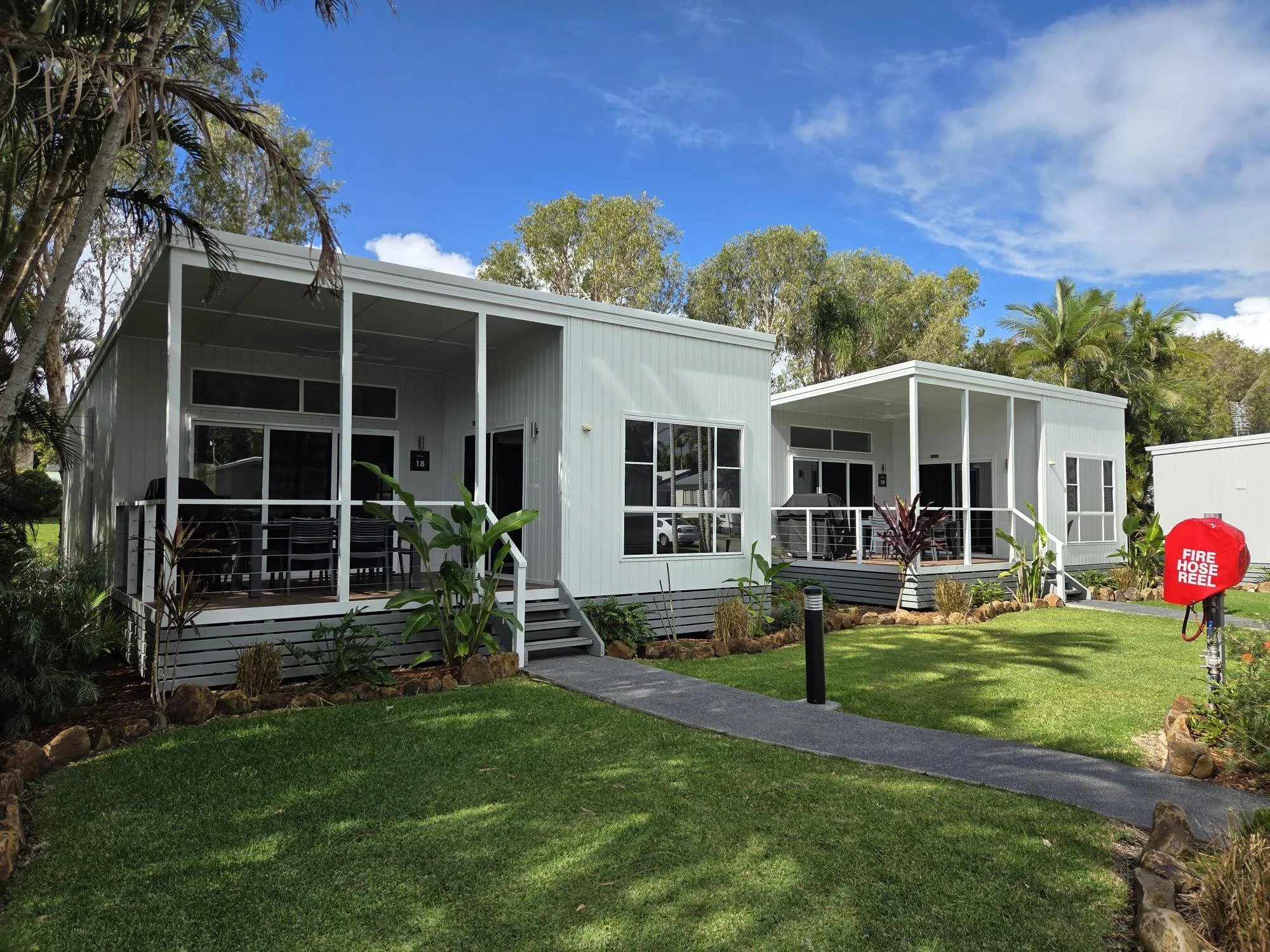 Two modern white houses with front porches, surrounded by a well-maintained lawn and tropical plants, under a blue sky with some clouds.