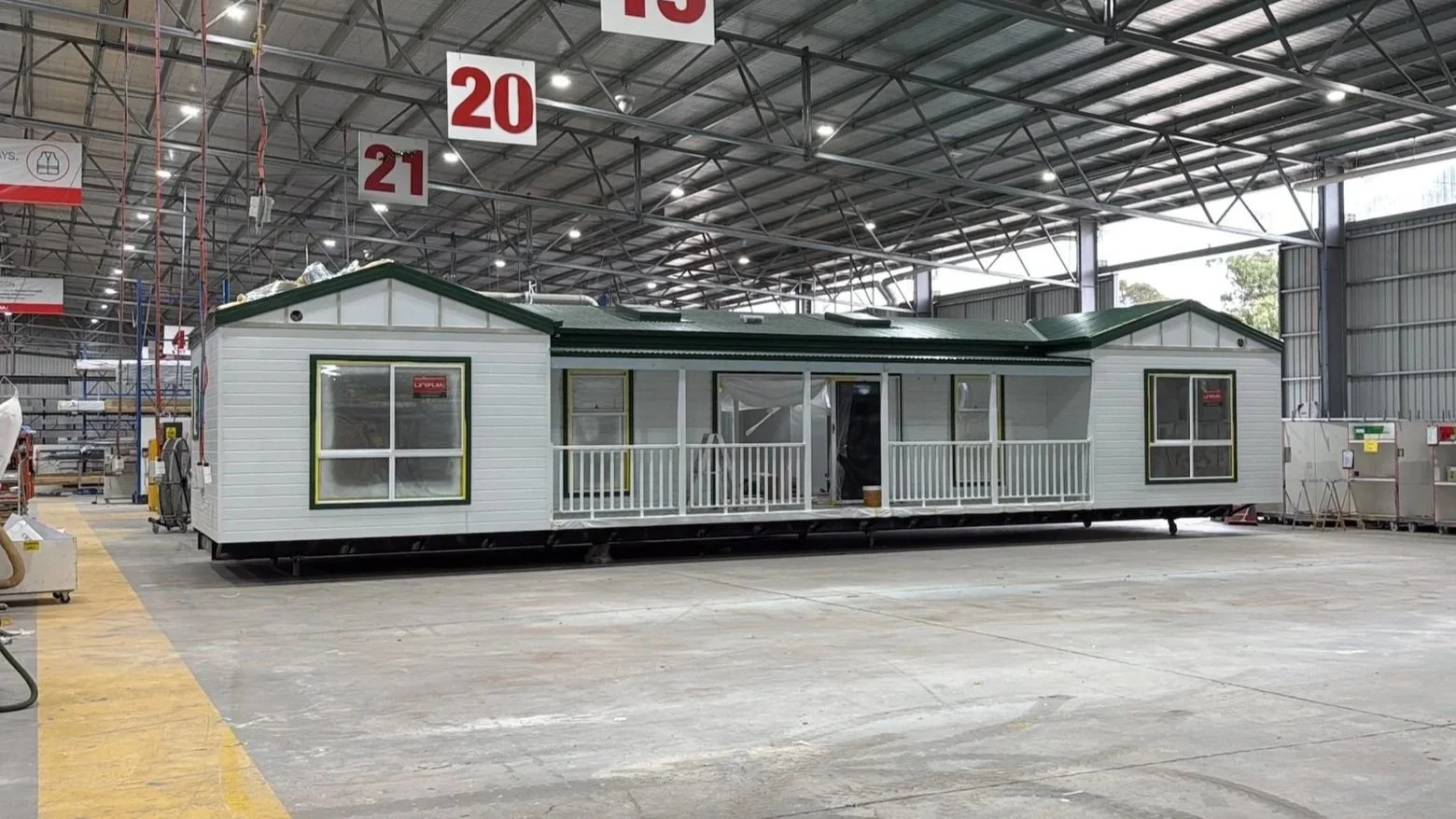 A manufactured tiny house with a porch and white siding, inside a large warehouse with signs overhead, including numbers 20 and 21.