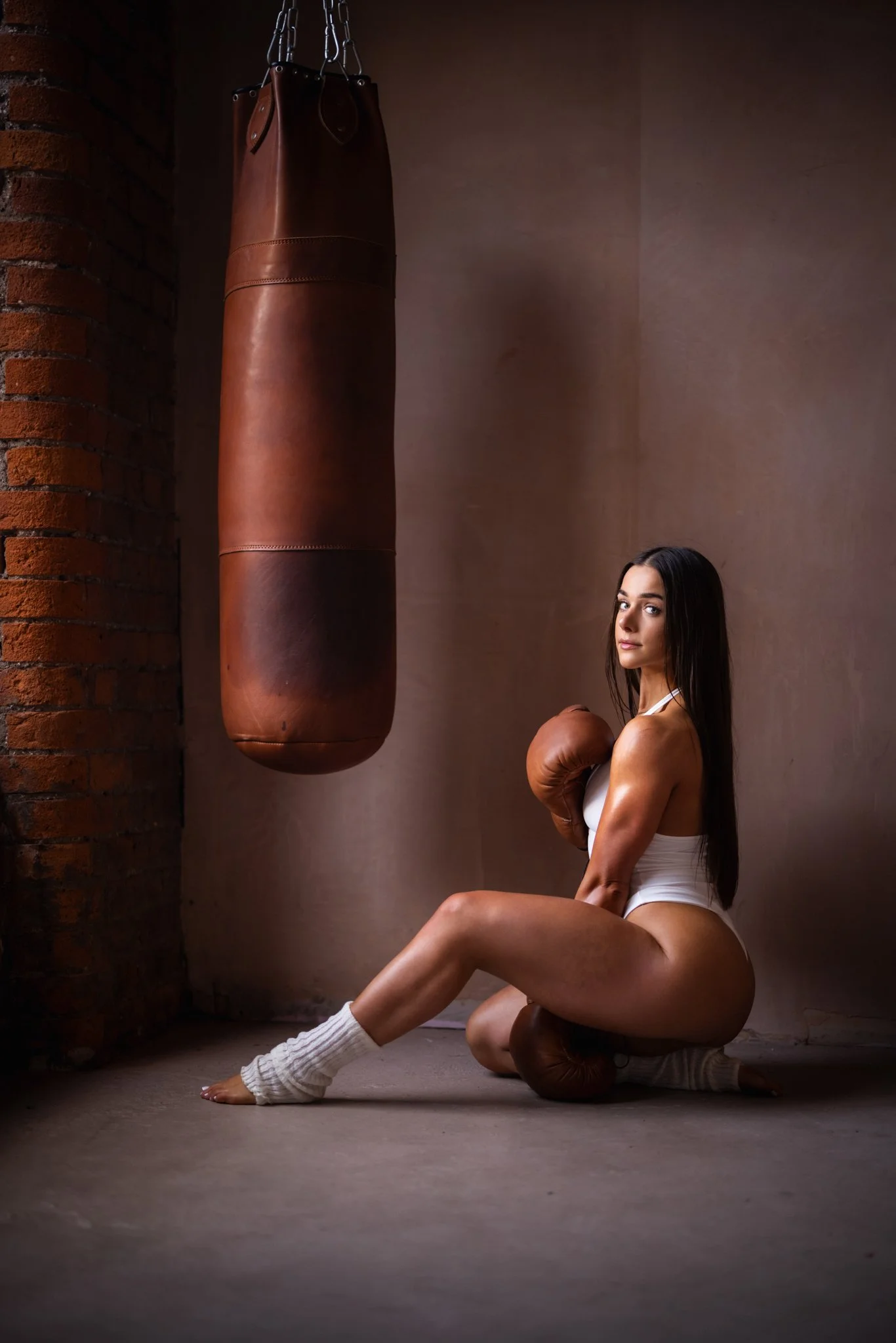 A woman in white sportswear with boxing gloves sitting on the floor near a hanging punching bag in a gym.