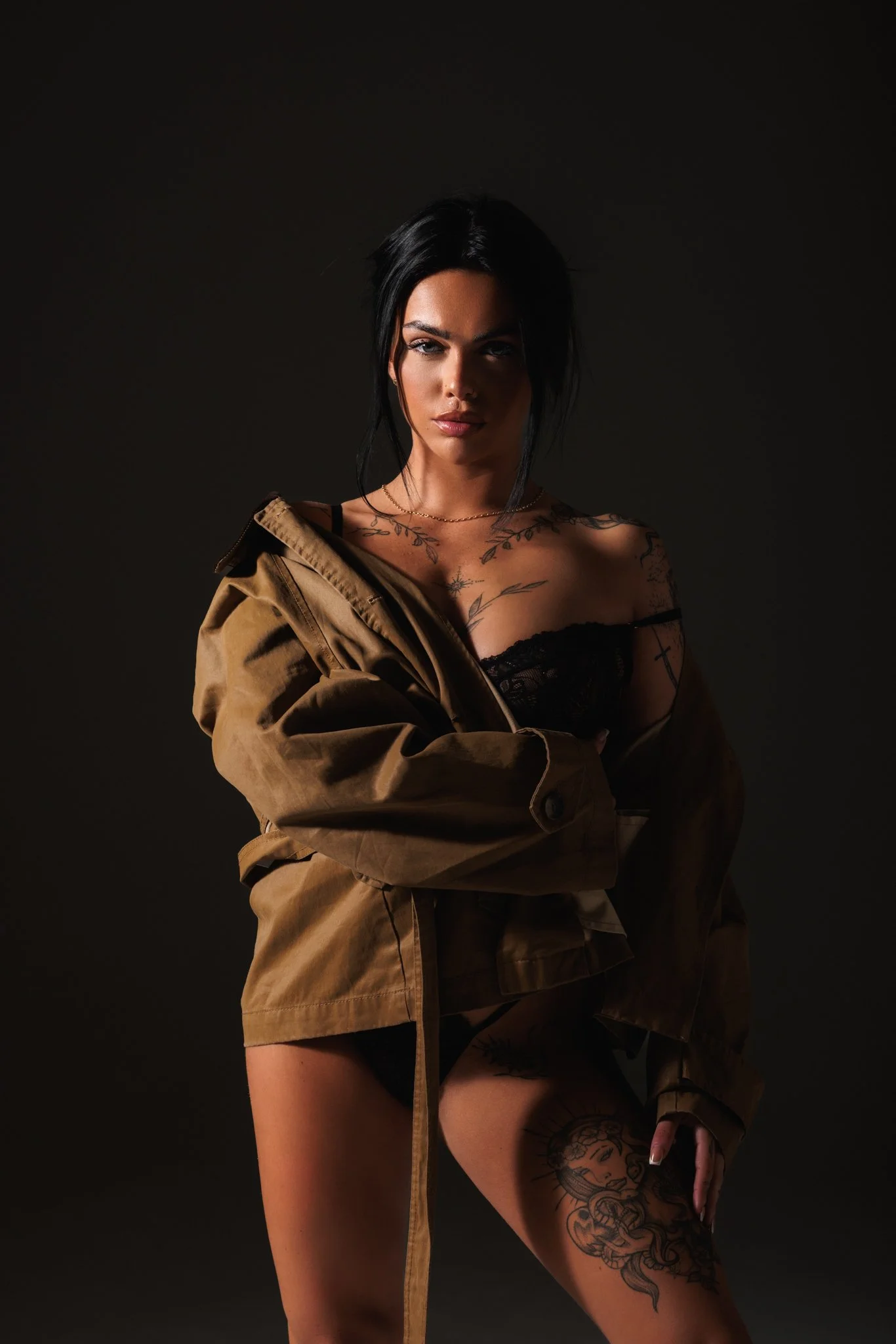A woman with dark hair and tattoos wearing a black lace top and tan jacket, posing against a dark background.