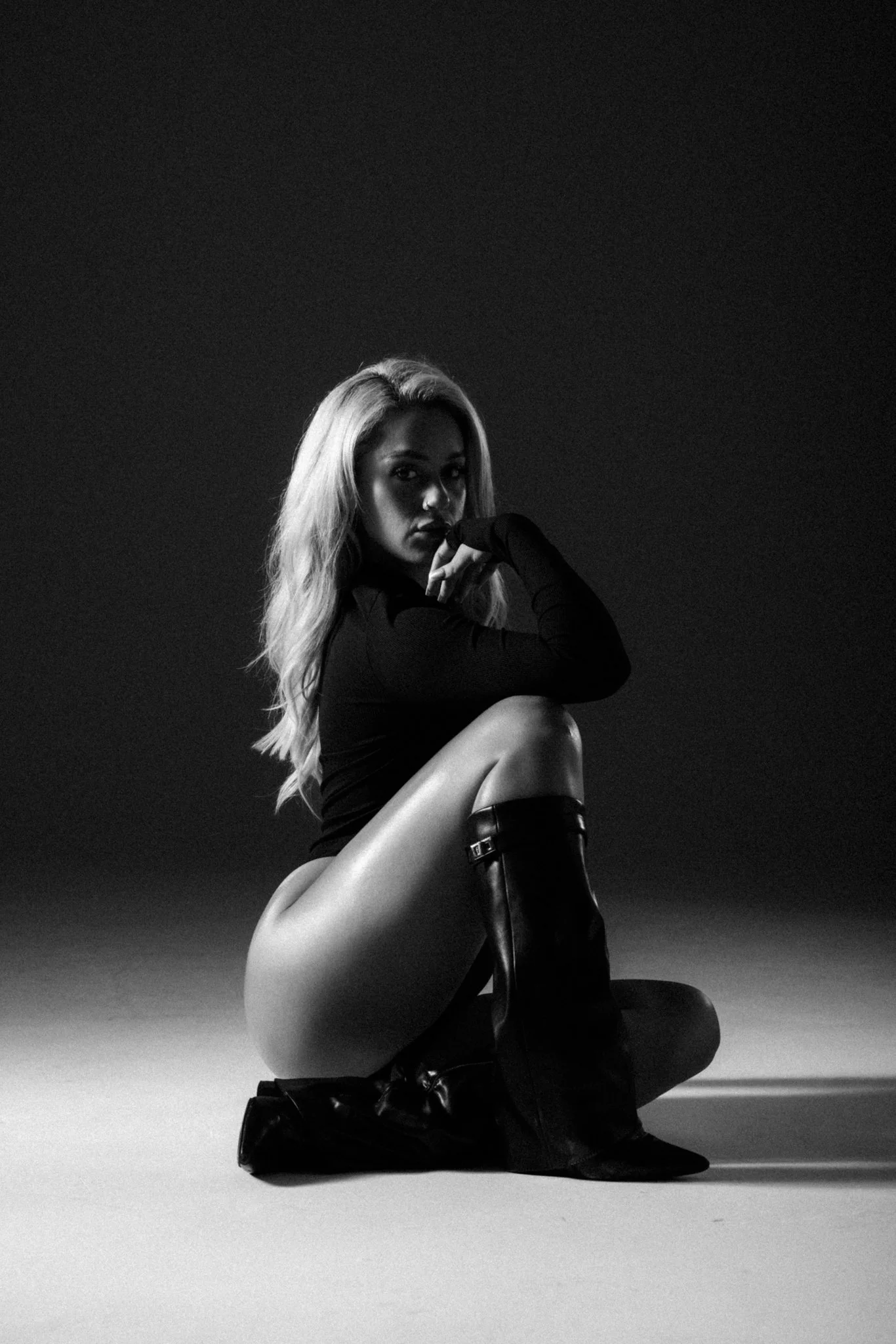 A black and white photograph of a woman with long blonde hair, kneeling on the ground with one knee bent and resting her chin on her hand. She is wearing a long-sleeved top, thigh-high boots, and the background is plain and dark.