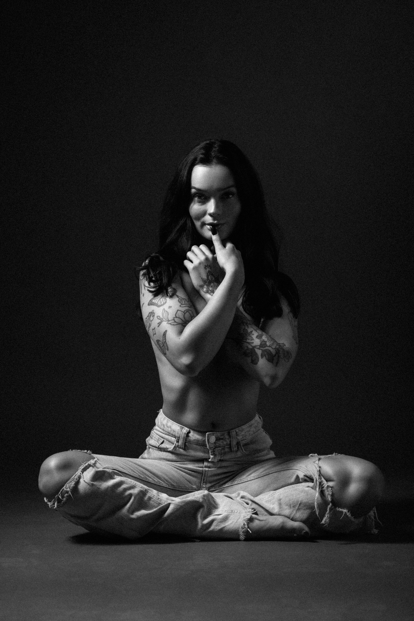 A young woman with dark hair, tattoos on her arms, sitting cross-legged in ripped jeans, holding a finger to her lips, posing in front of a dark background.