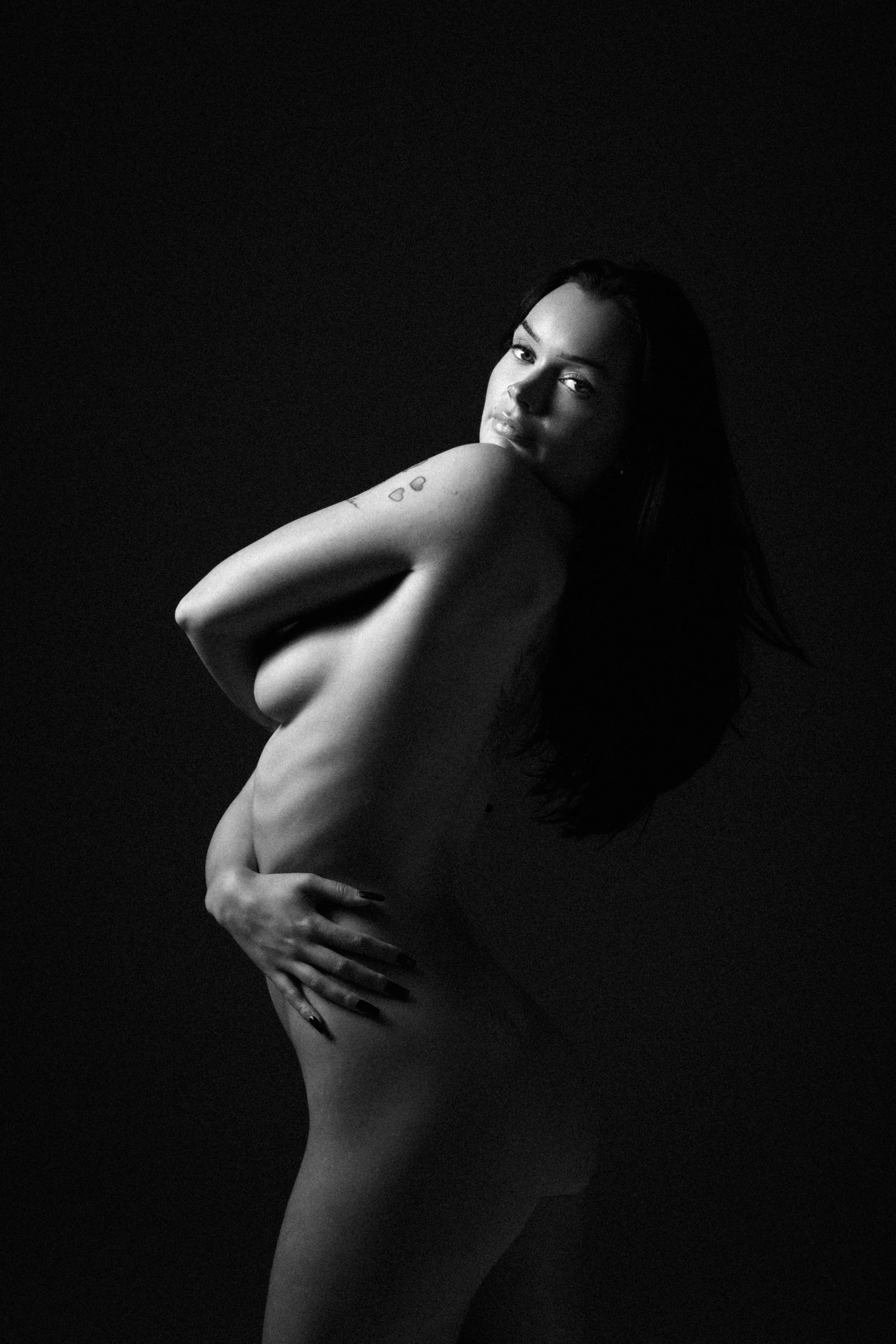 A black and white photograph of a nude woman with long dark hair posing against a plain background. She is looking at the camera with her body slightly turned to the side, one arm covering her chest, and the other hand on her hip, with small tattoo h