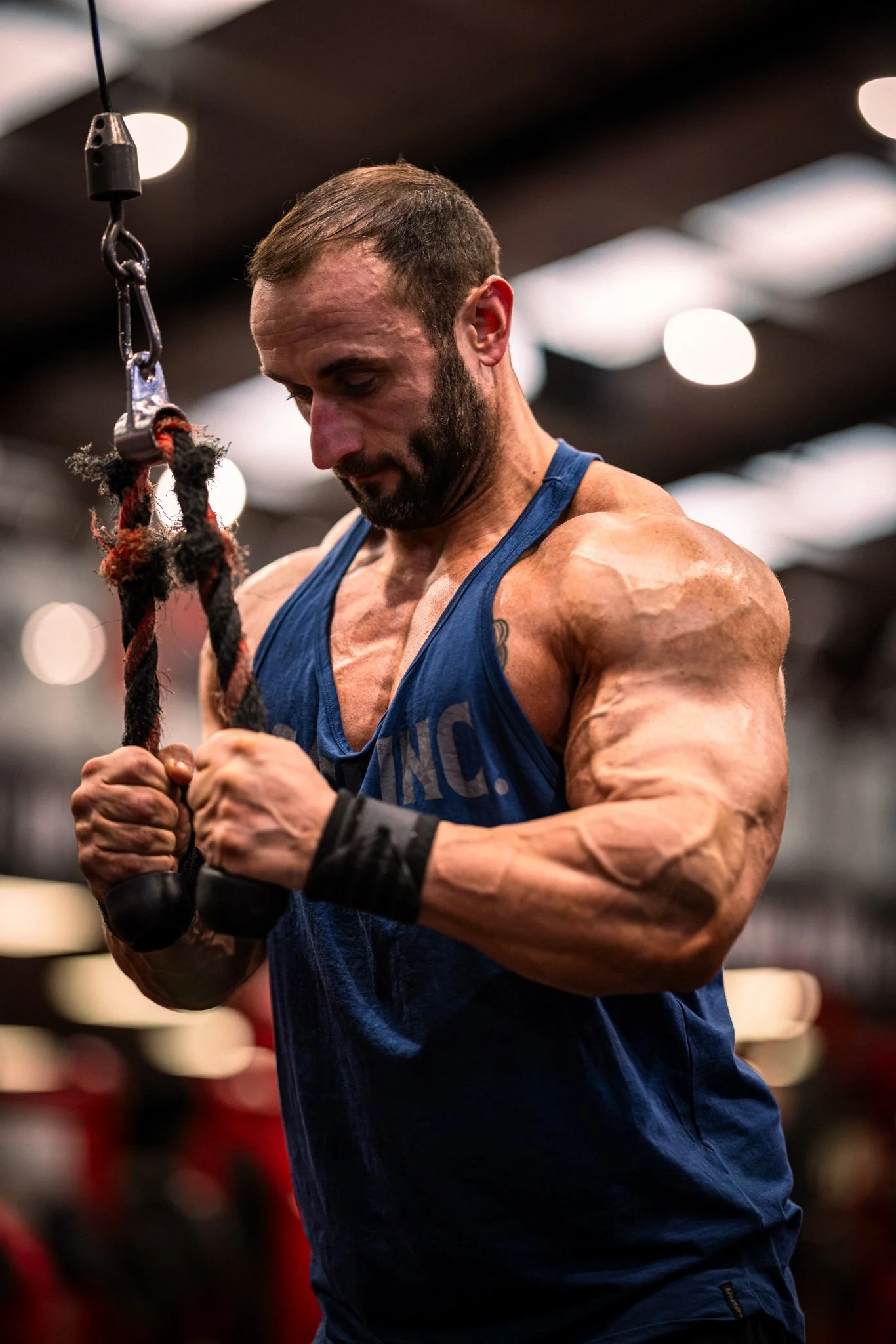 A muscular man in a blue sleeveless shirt is working out in a gym, gripping a rope attachment for strength training.