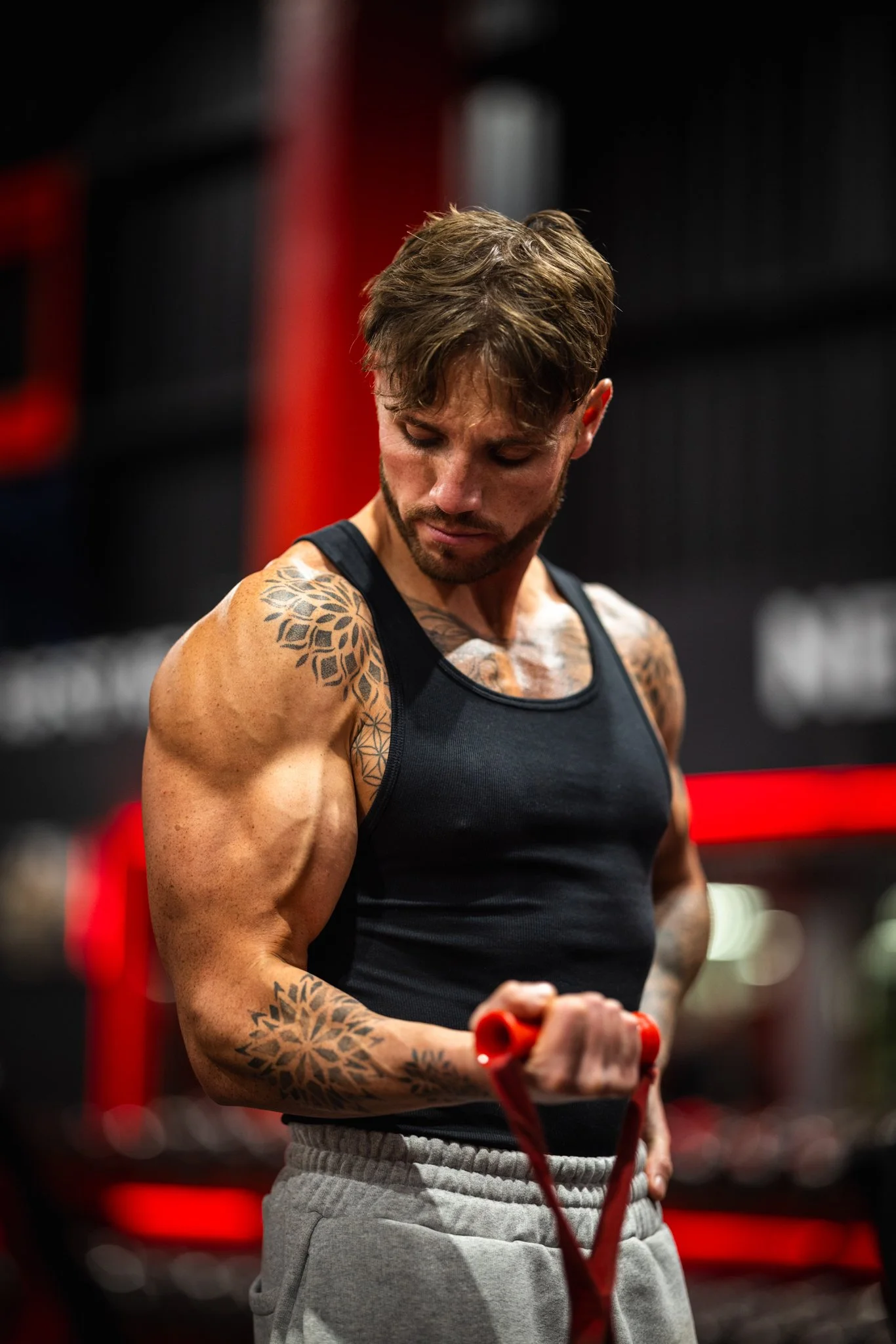 A muscular man with tattoos in a black tank top and gray sweatpants is working out with a resistance band in a gym.