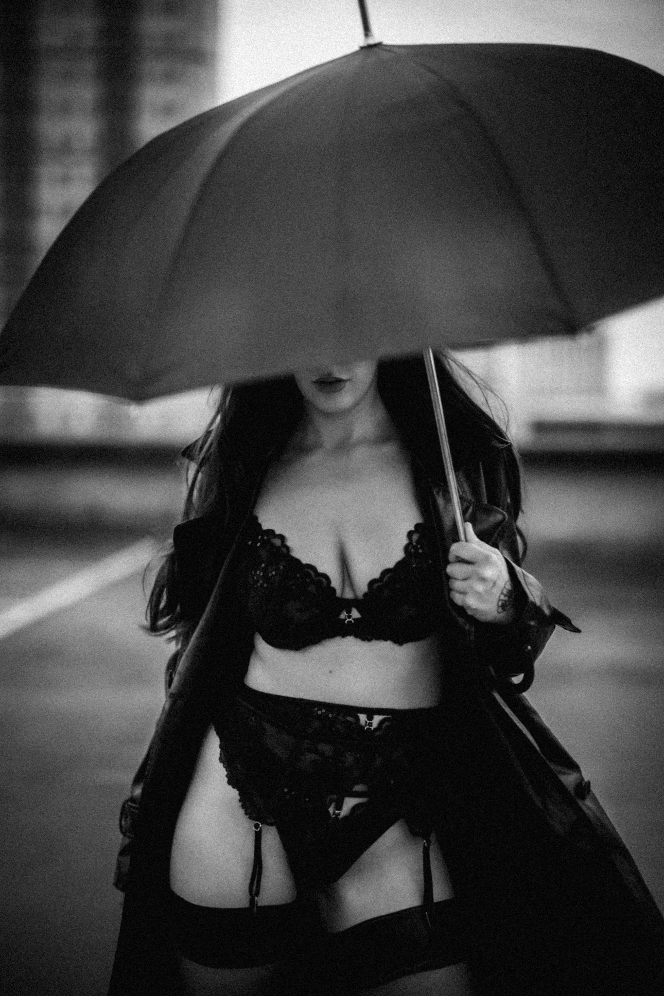 A woman holding an umbrella, wearing black lace lingerie and a leather jacket, walking on a street at night.