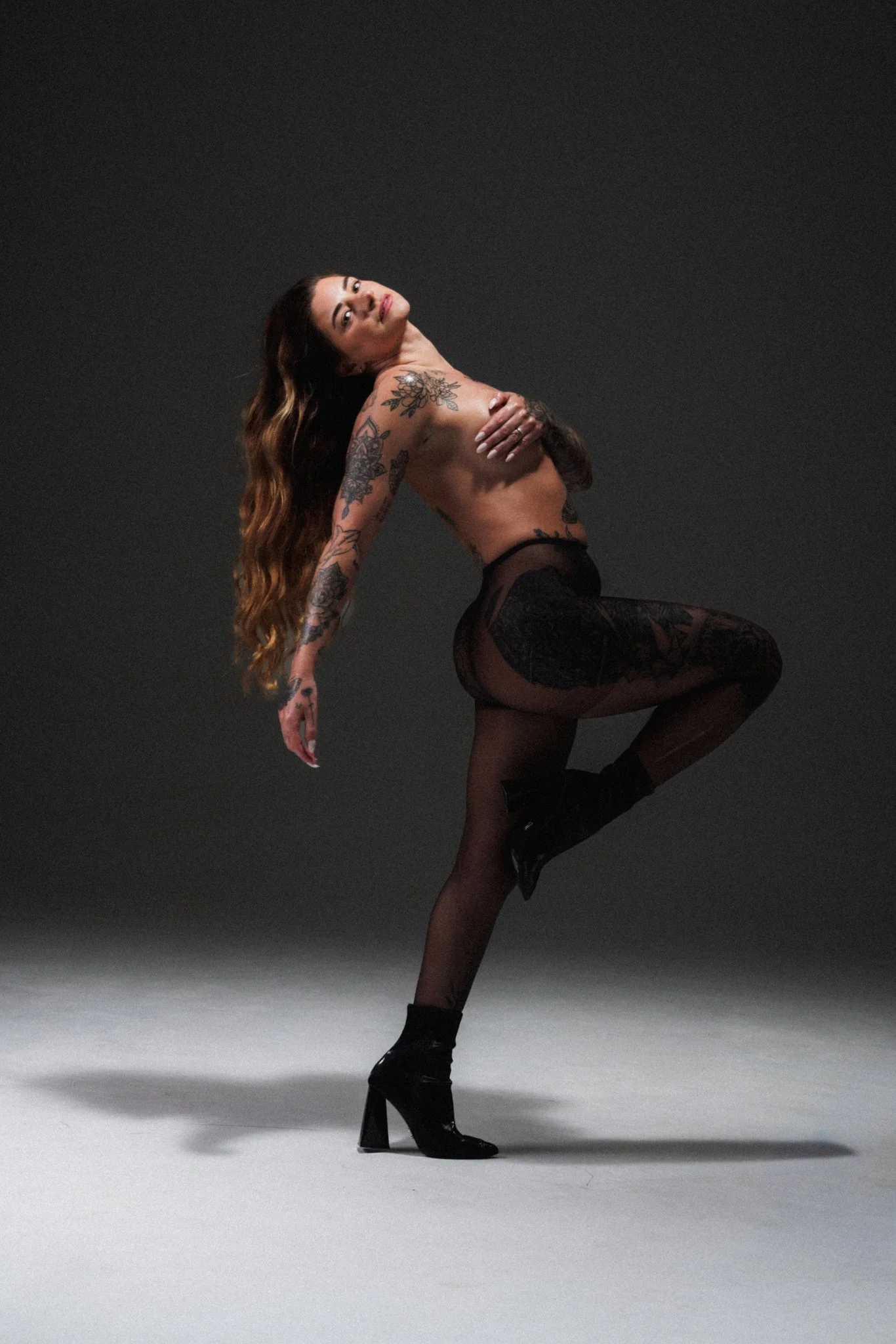 A woman with long wavy hair and tattoos on her arms and torso poses in black stockings and high heels against a gray backdrop, with one knee bent and her hand covering her chest.