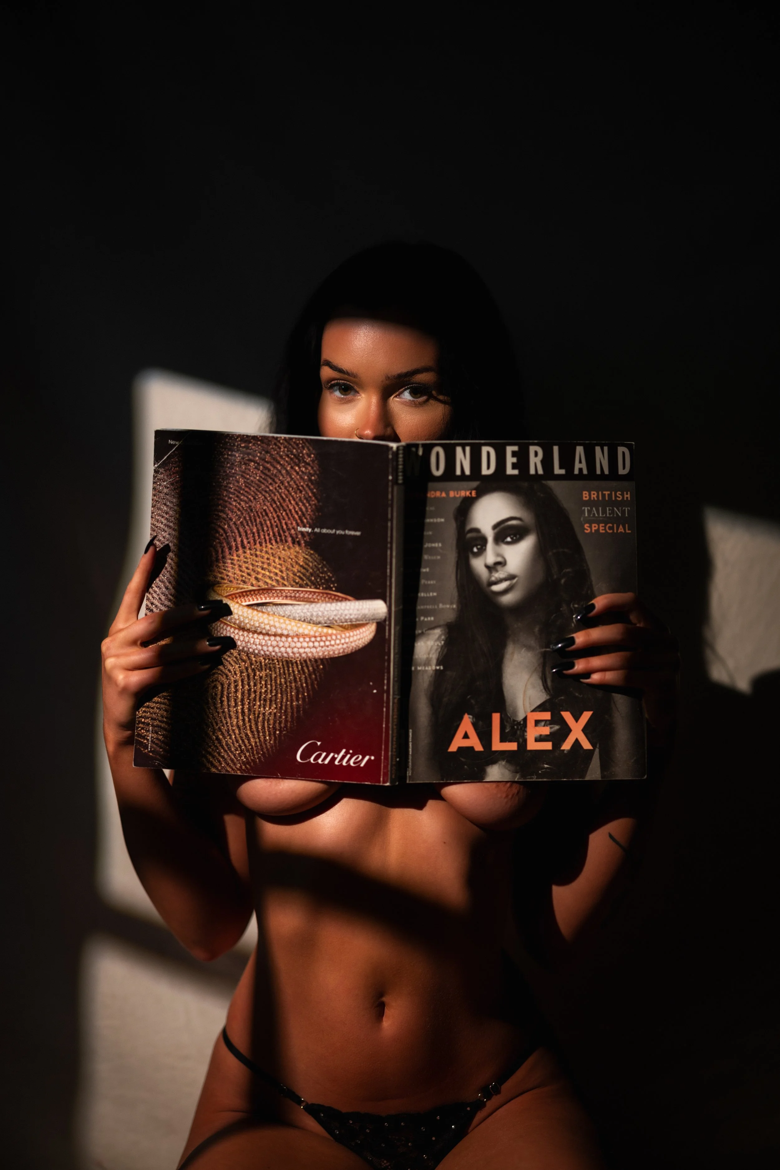 A woman with dark hair and black lingerie holding a magazine covering her face and chest, with light and shadow effects in a dark room.