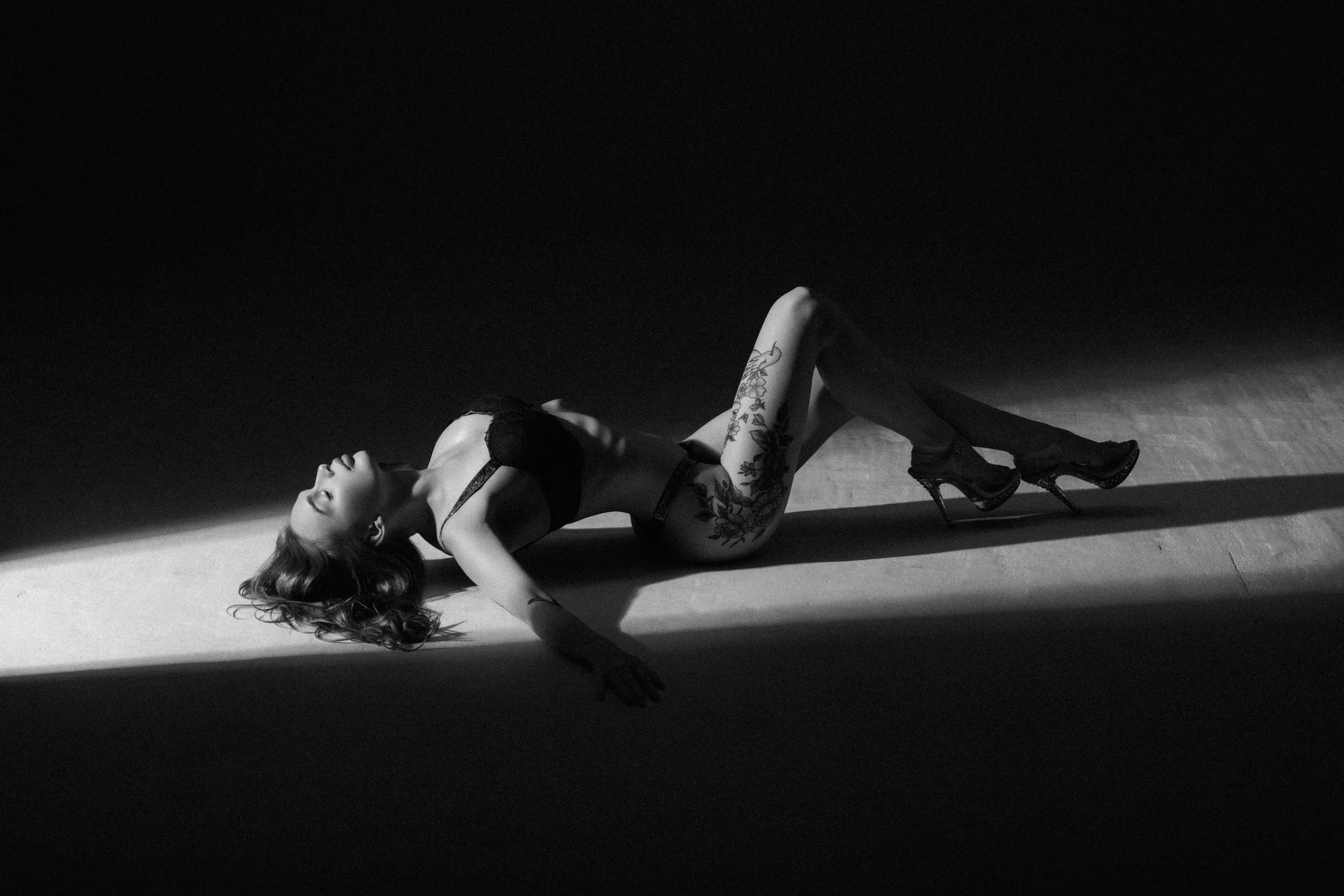 A woman lying on the floor in a black bra and stockings, with her eyes closed and head tilted back. She is illuminated by a strong light creating a shadow on the floor, highlighting her tattoos and high heels.