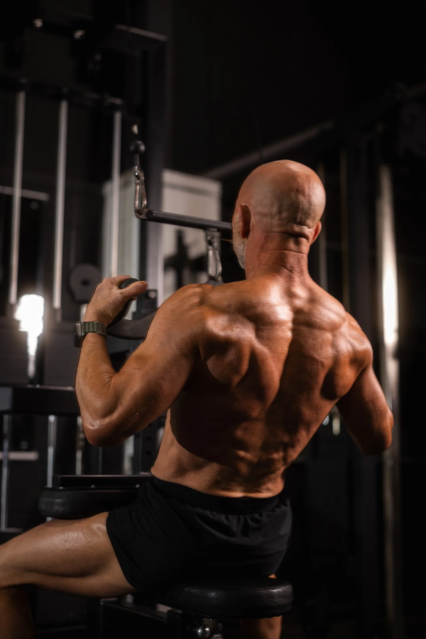 A muscular, bald man with a beard sitting on a bench and performing a shoulder exercise in a gym, using a machine with metal bars and pulleys.