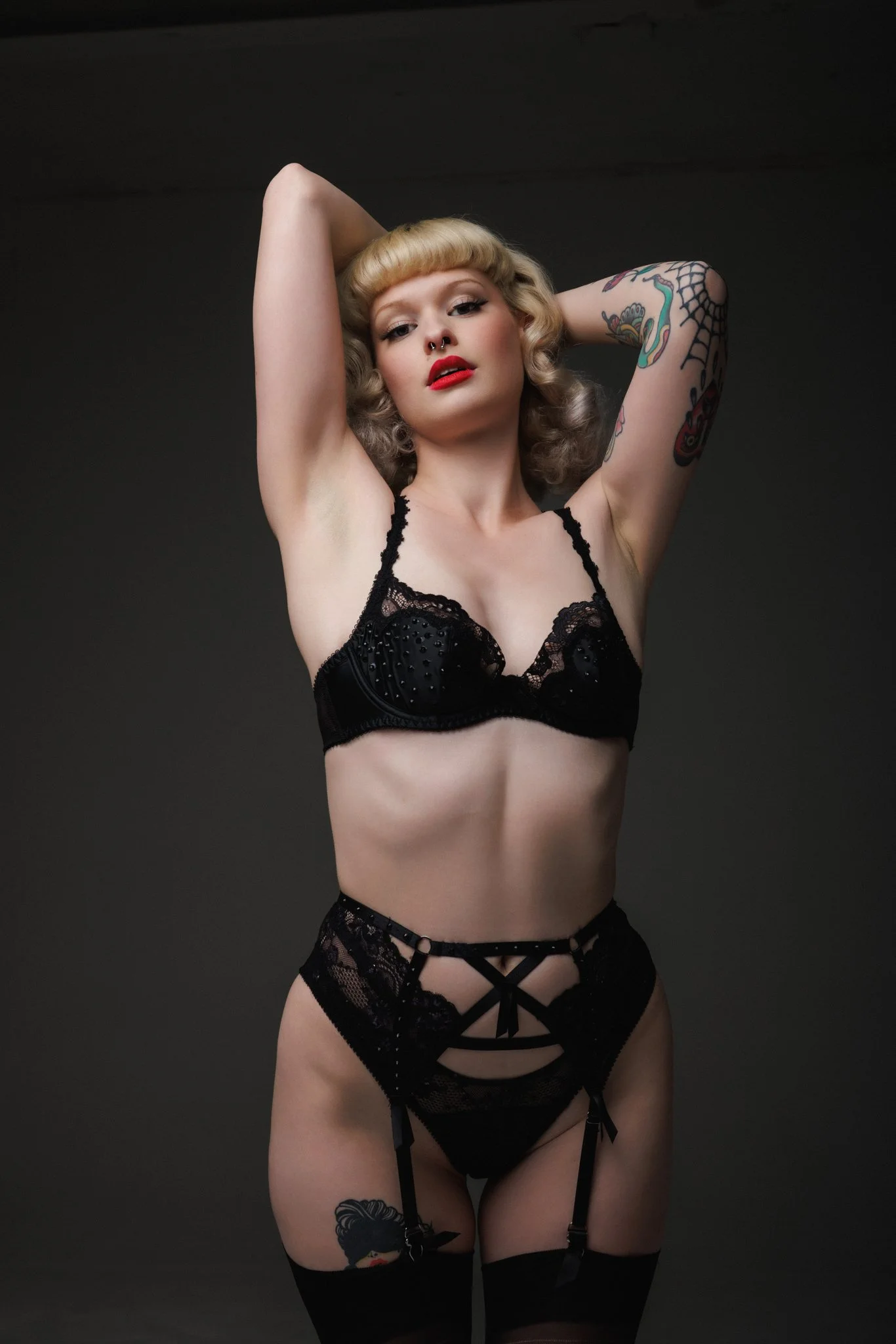 A woman with tattoos and blonde hair wearing black lingerie and thigh-high stockings, posing with arms behind her head against a dark background.