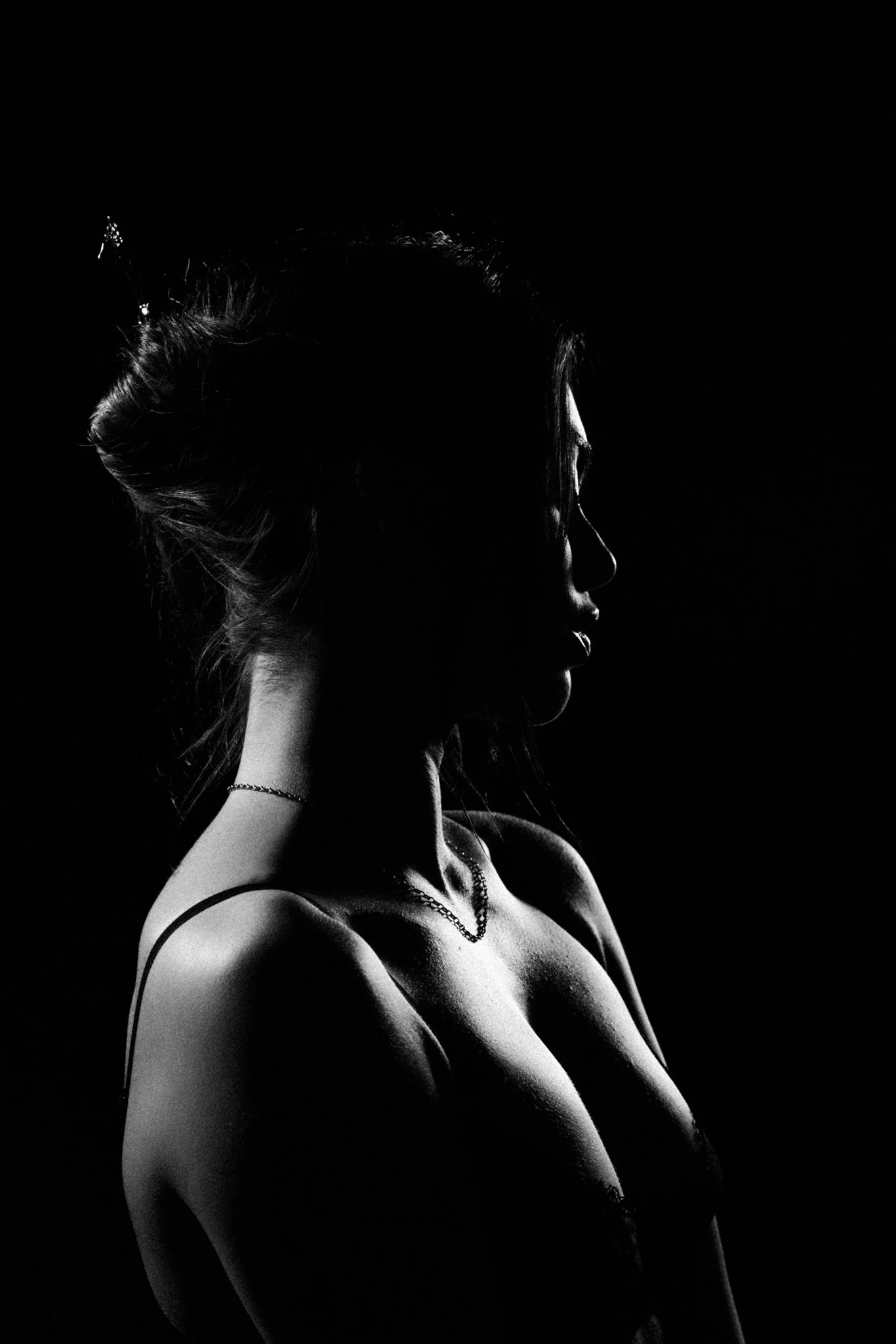 Silhouette of a woman with her head turned to the side, illuminated from the side, showing her profile, with her hair in a bun and wearing a tank top and a necklace.