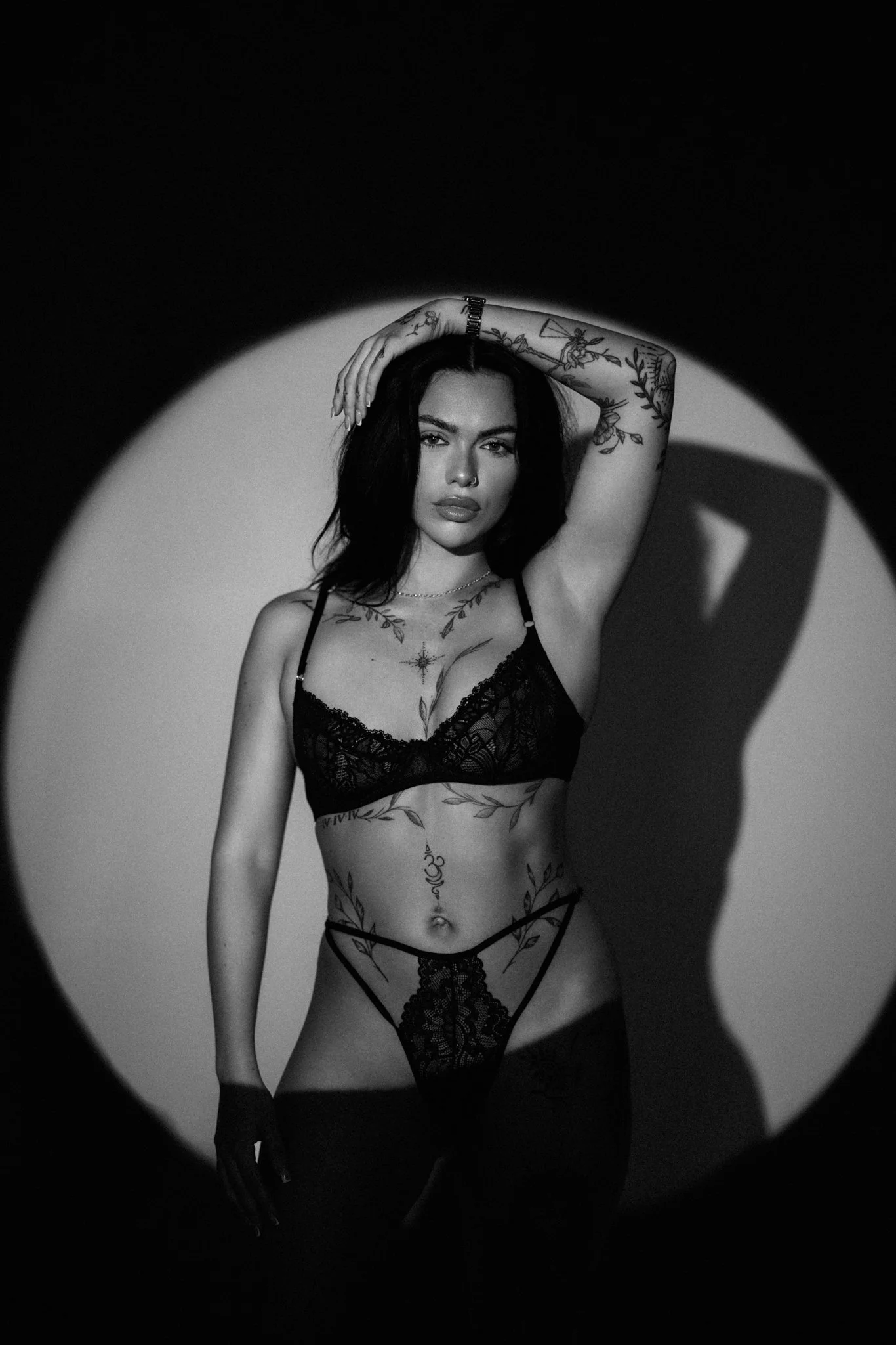 A woman with tattoos wearing black lingerie, standing in front of a spotlight creating a circular shadow behind her.
