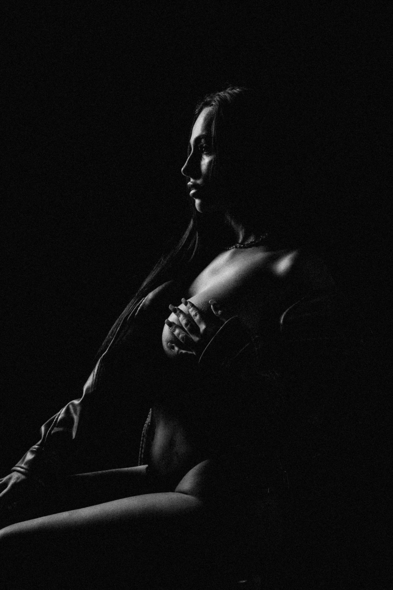 Black and white portrait of a woman, lit from the side, with her hand resting on her chest. Her face and upper body are visible, with shadows creating a dramatic effect.