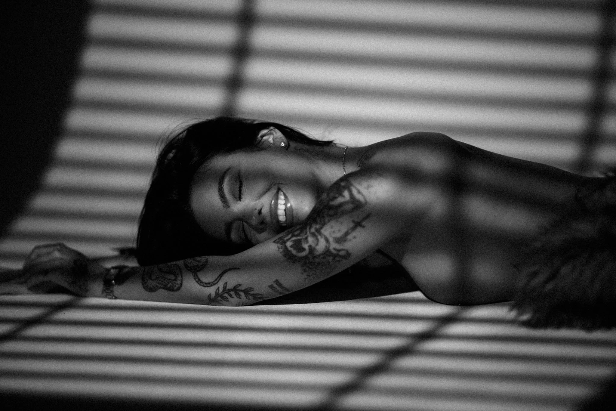 A woman with dark hair and tattoos smiling and lying down, viewed through horizontal blinds.