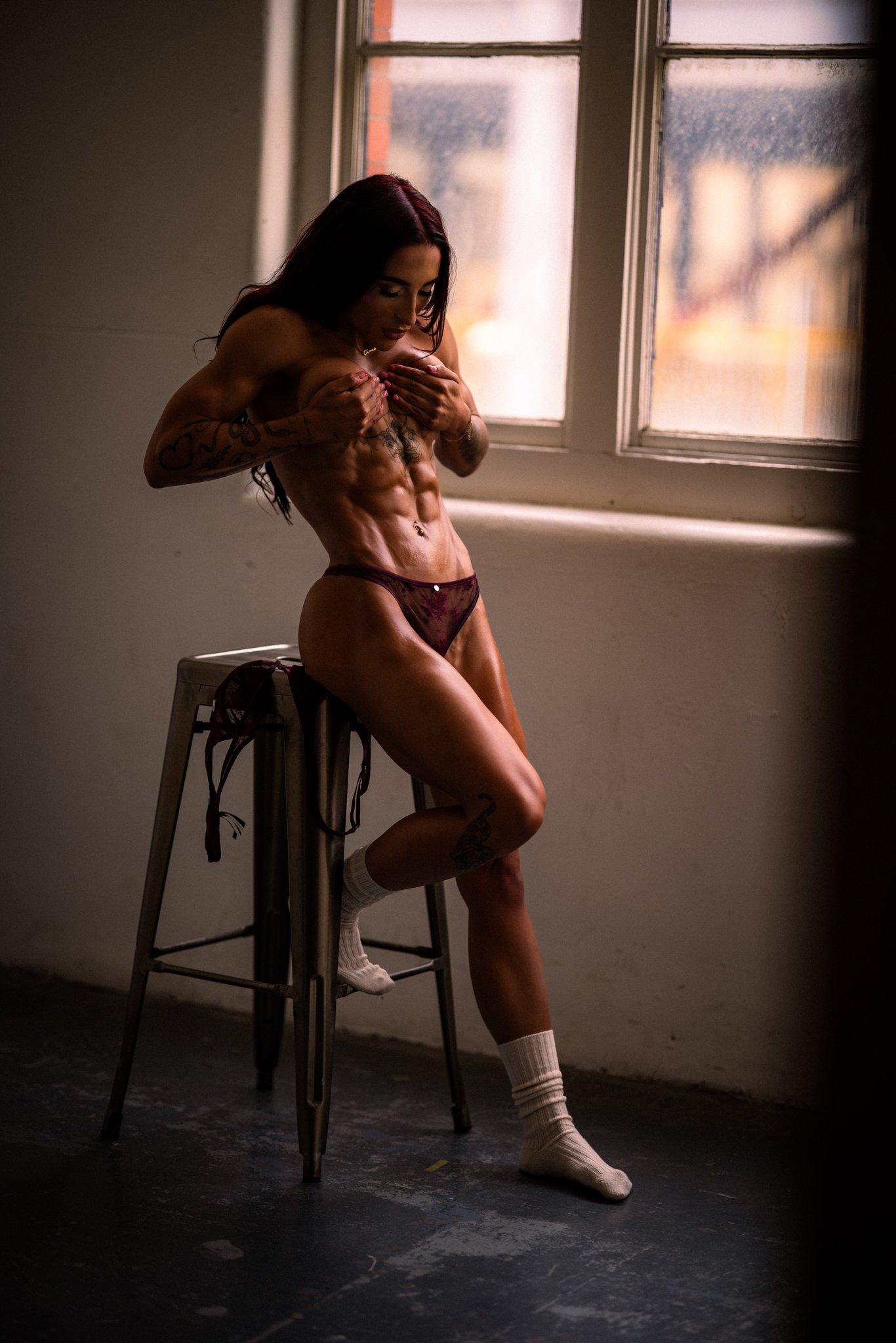 A woman with tattoos, wearing burgundy underwear and white socks, sitting on a stool near a window with her hands covering her chest, looking down.