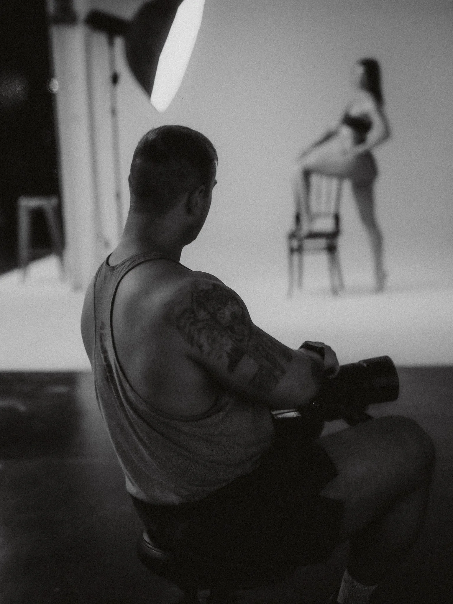A man with tattoos on his arm sitting on a stool, facing a woman who is balancing on a chair, in a photography studio setting.