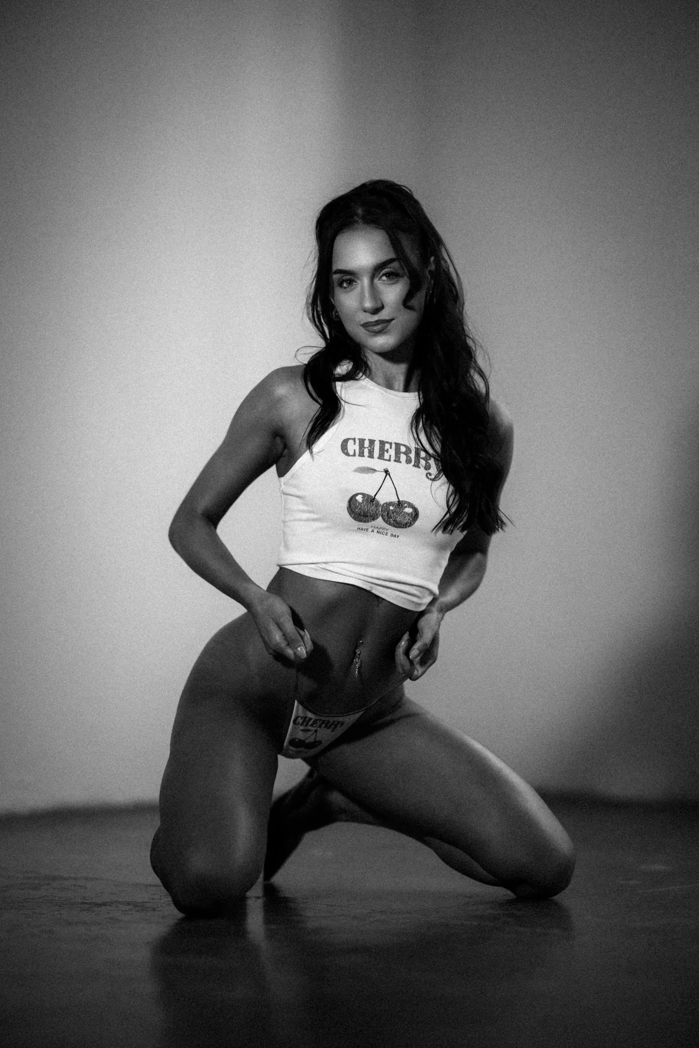 A black and white photo of a woman with long dark hair, wearing a sleeveless cherry print shirt, kneeling on the floor, looking at the camera with a confident expression.
