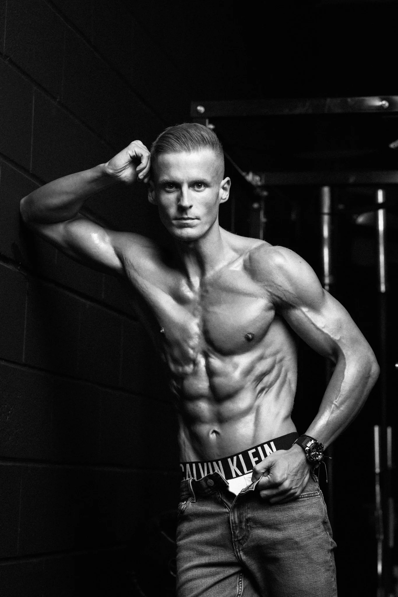 A shirtless man with a muscular physique, short hair, and wearing jeans with Calvin Klein waistband visible, poses against a dark background.