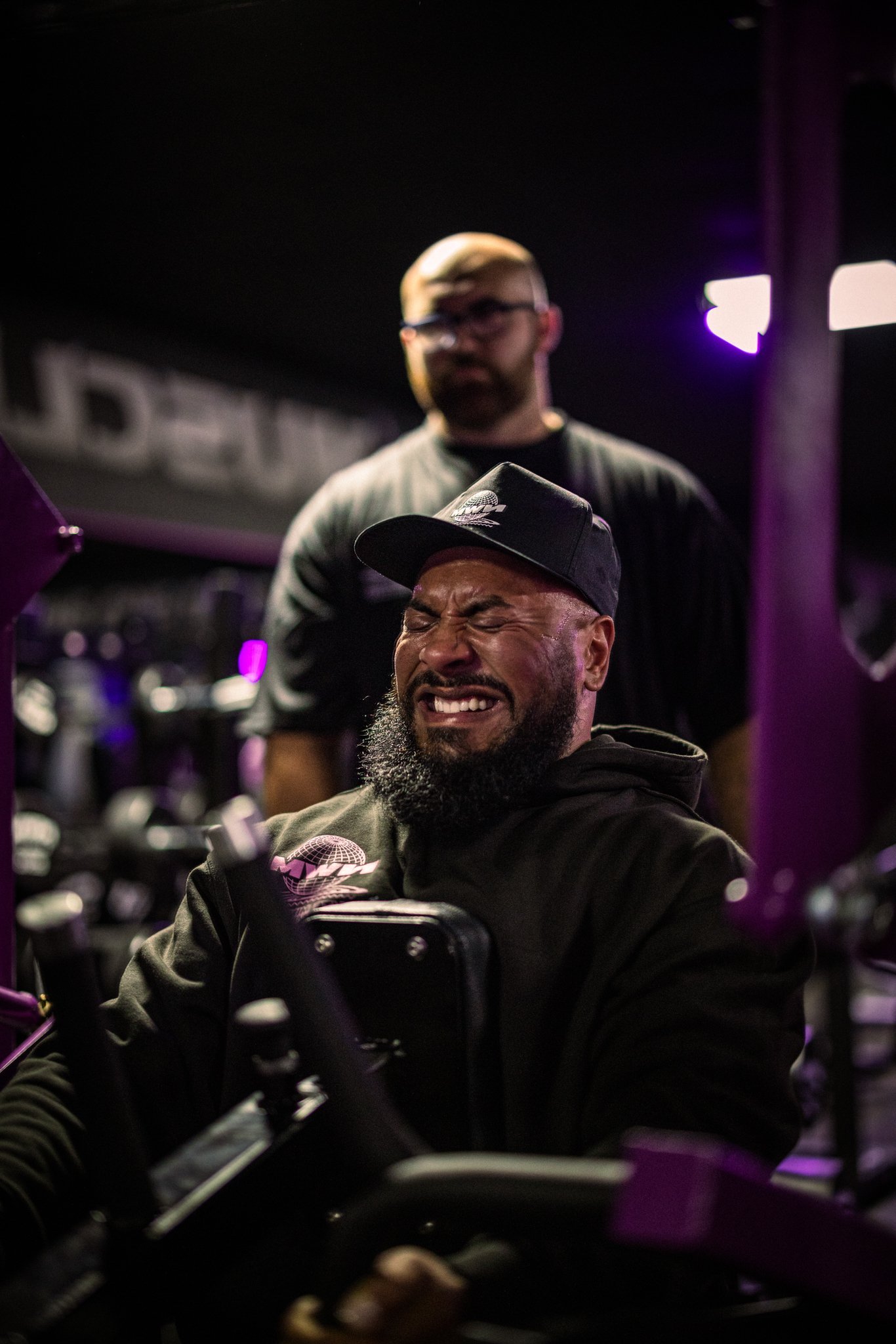 A man with a beard and black cap grimacing while riding a motorcycle, with a second man standing behind him, in a dimly lit indoor setting with purple lighting.