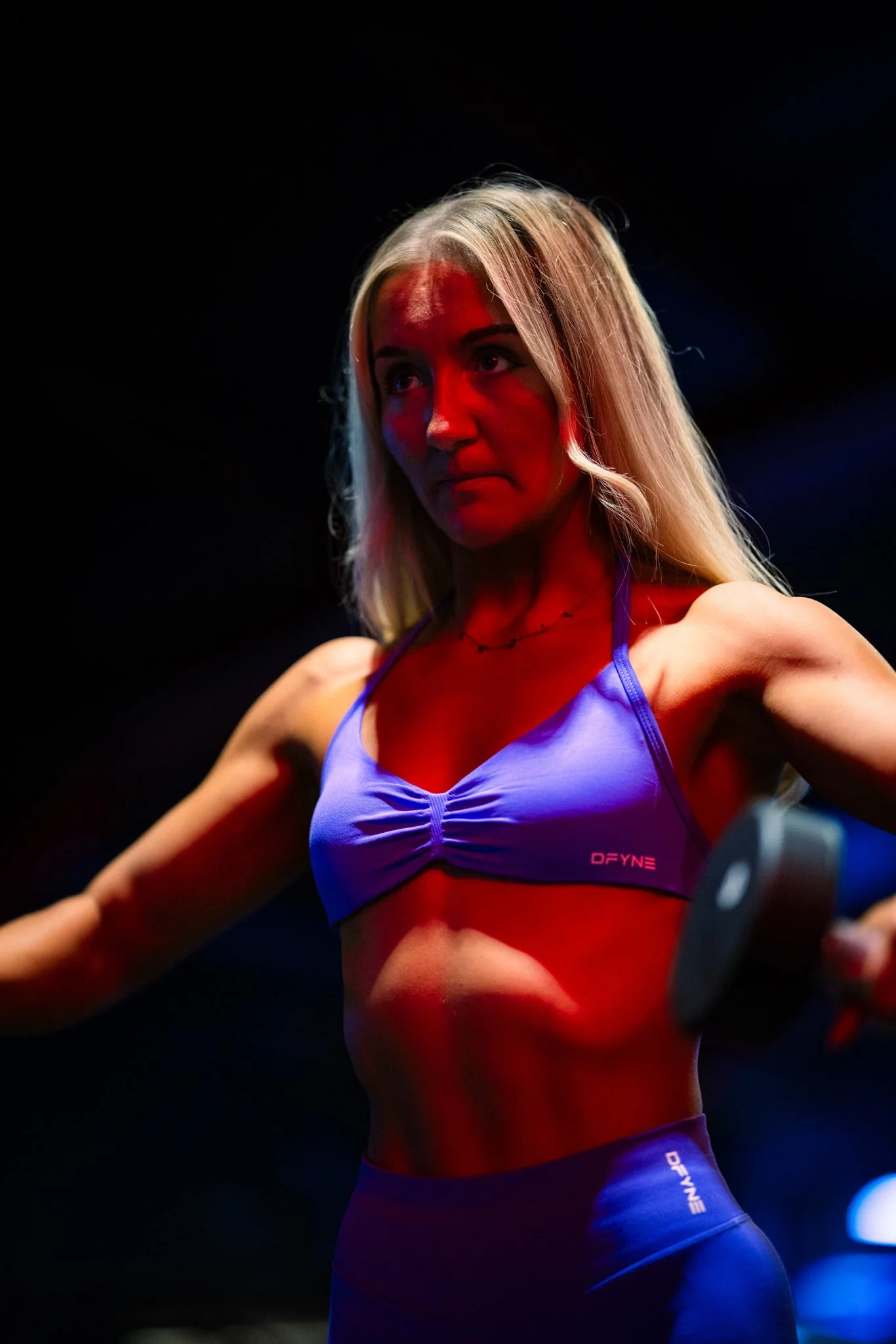 A woman with long blonde hair exercising with dumbbells in a dark gym illuminated by red and blue lights.