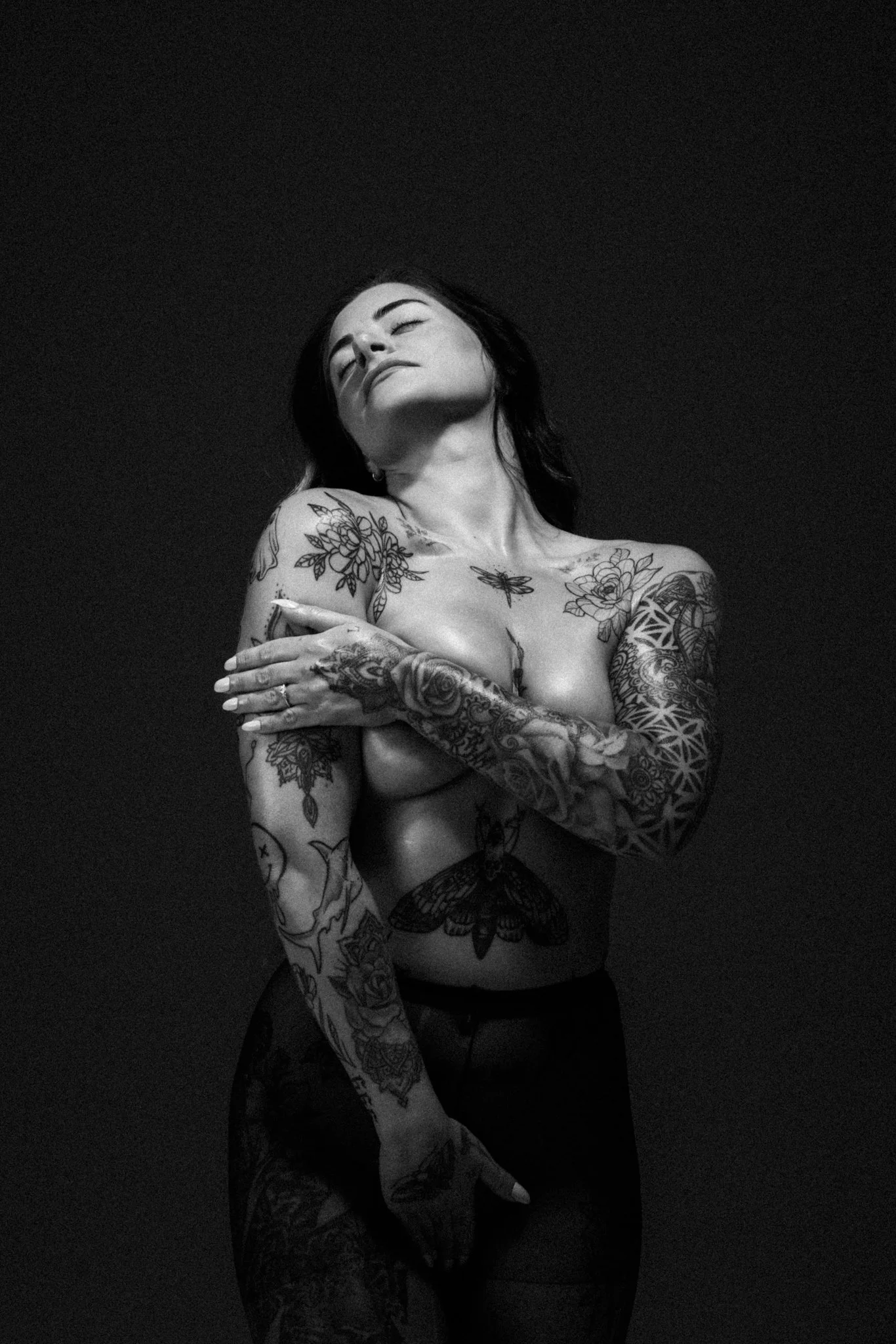 A black-and-white photo of a woman with long dark hair and numerous tattoos on her arms, shoulders, and chest. She is topless, with a serene expression on her face and eyes closed, against a dark background.