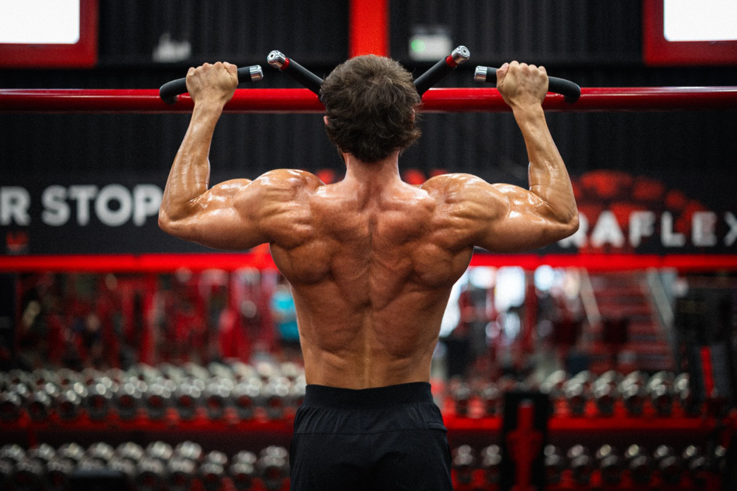 A muscular man with bare upper body lifting a pull-up bar in a gym with red and black equipment in the background.