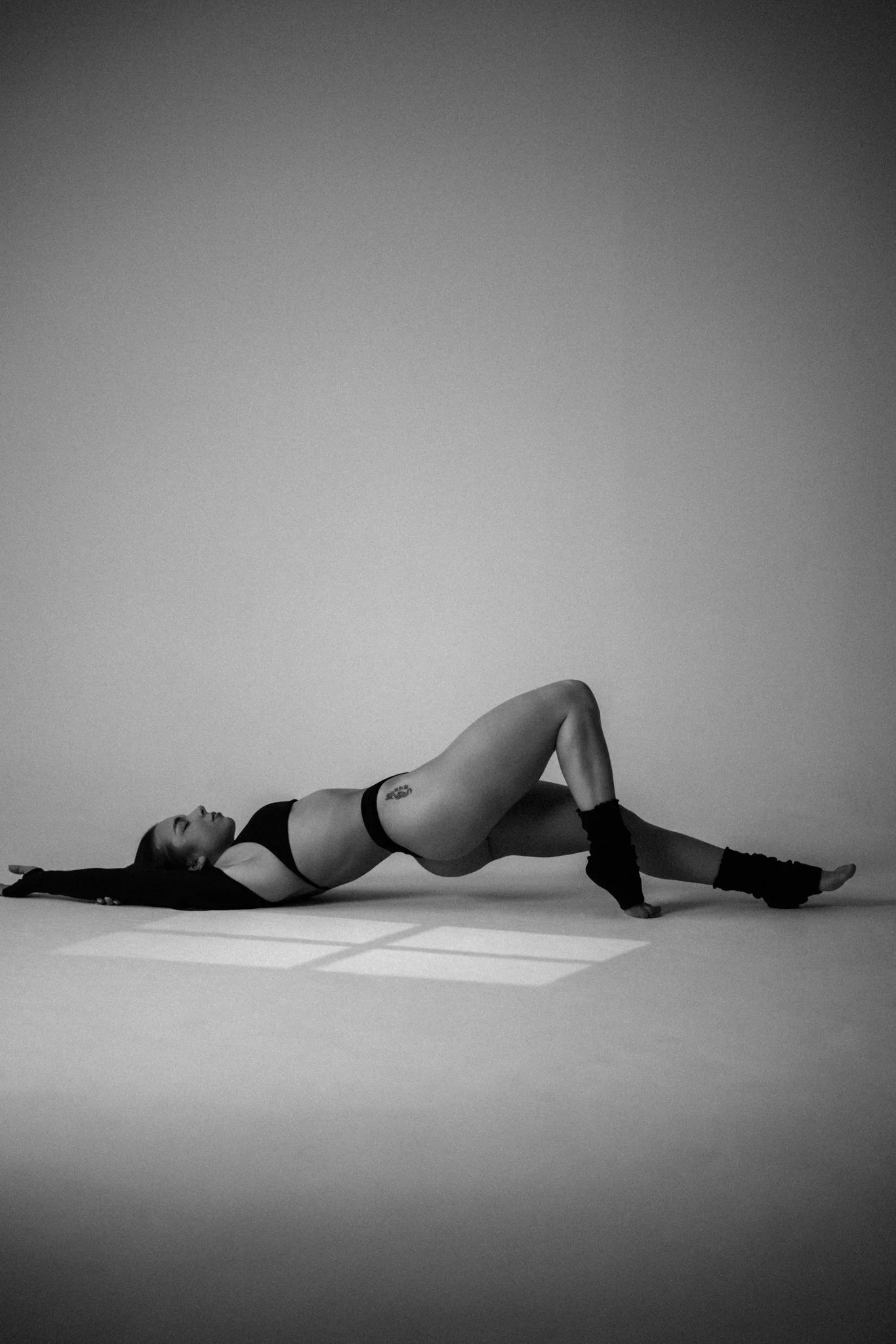 A woman lying on her side on the floor, supporting herself with one arm and a leg raised, wearing a black sports bra, high-waisted shorts, and leg warmers, against a plain background.