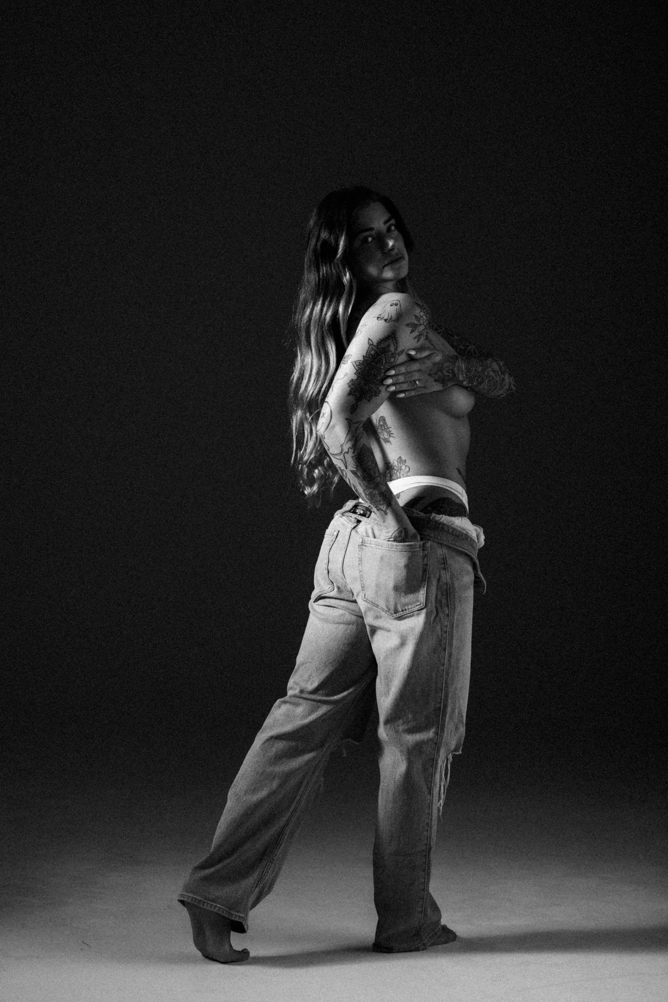 Black and white photo of a woman with long wavy hair, tattoos on her arms and torso, wearing baggy jeans and no shirt, standing with one foot on the ground and the other on her toes, looking over her shoulder at the camera in a studio setting.