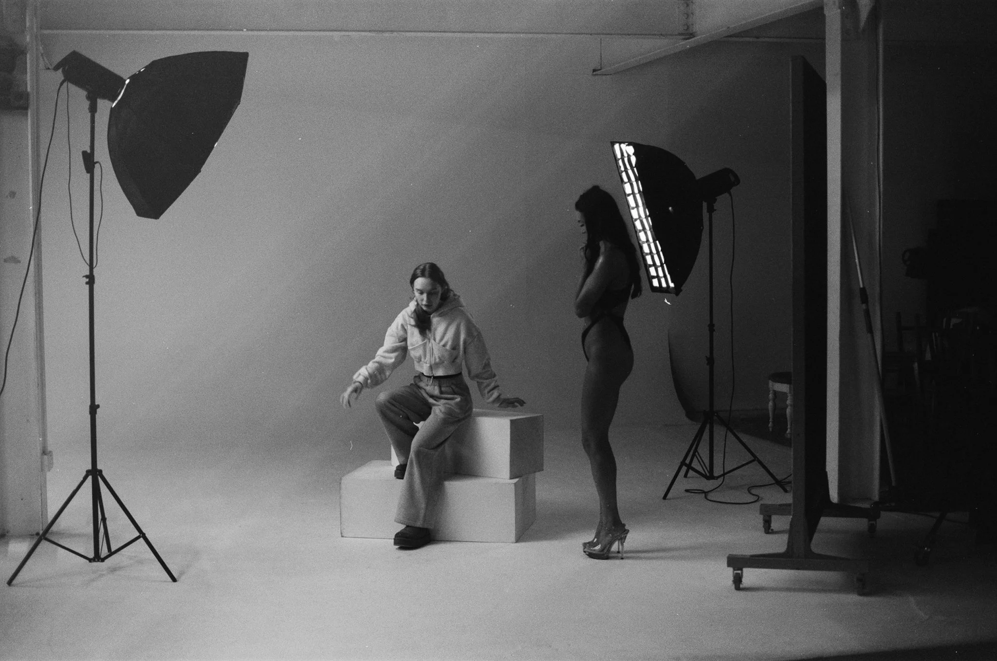 Two women in a photography studio with studio lighting equipment. One woman is seated on white blocks, wearing casual clothing. The other woman is standing, dressed in lingerie, and wearing high heels.
