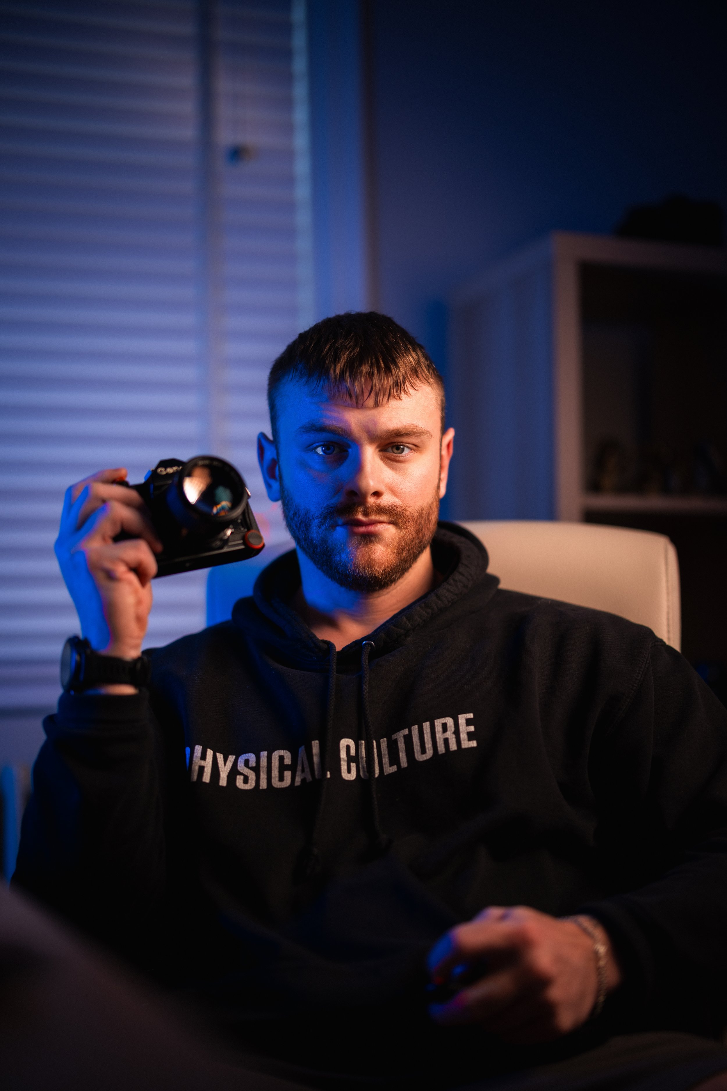 A man with short brown hair and a beard sitting in a room with blue and purple lighting, holding a camera in his right hand, wearing a black hoodie with white text that reads 'PHYSICAL CULTURE'.