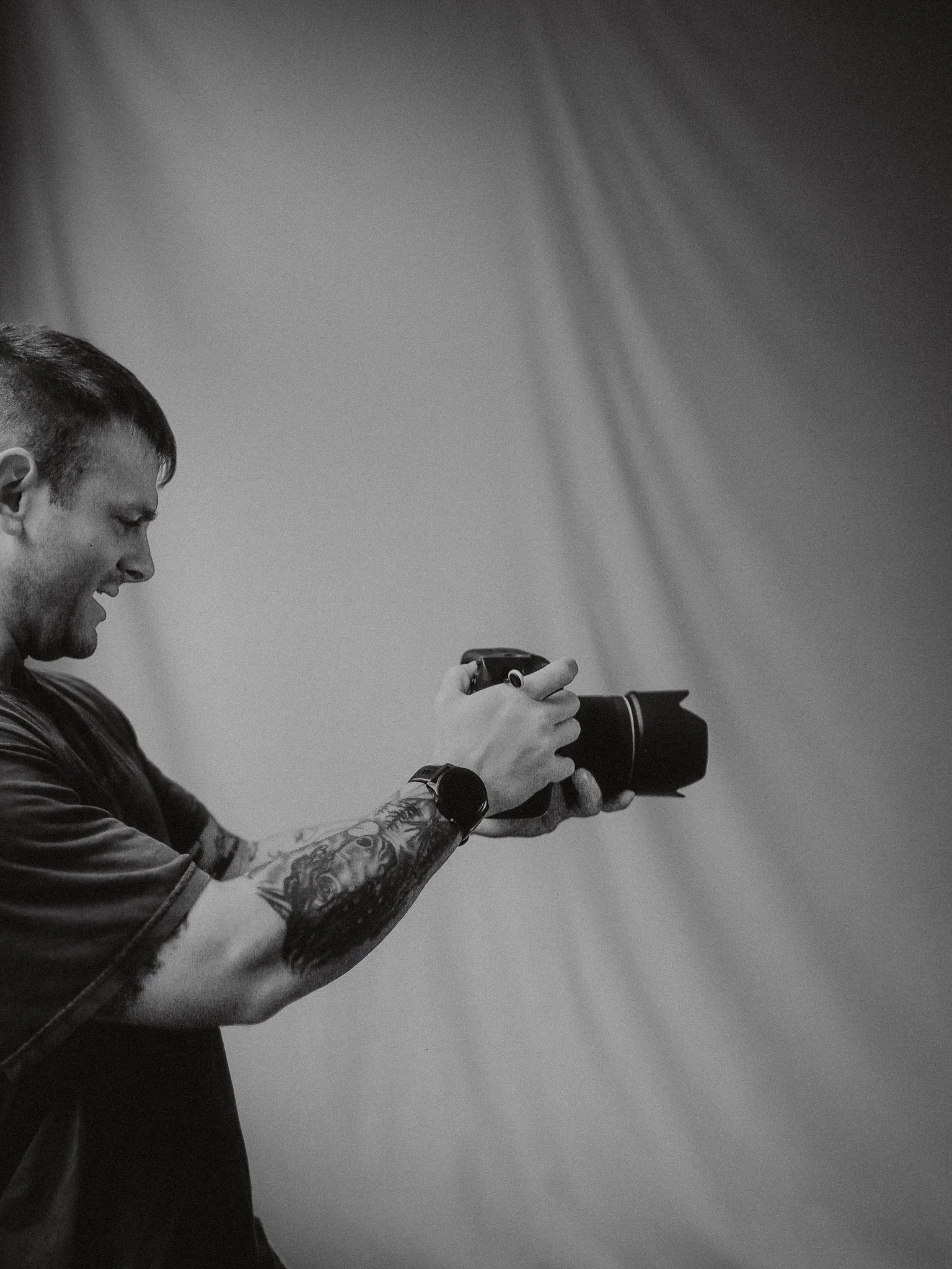 A young man with tattoos on his arm is smiling and taking a photograph with a DSLR camera against a plain wall.