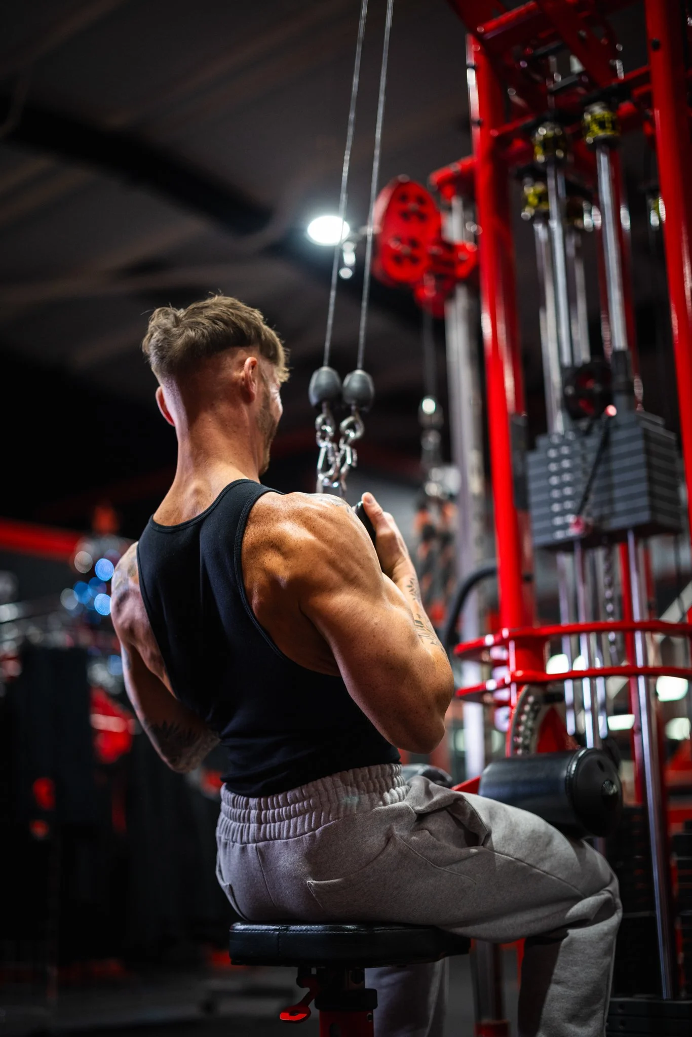 A muscular man with tattoos in a black tank top and gray sweatpants sitting on a workout bench and using a cable machine for strength training in a gym.