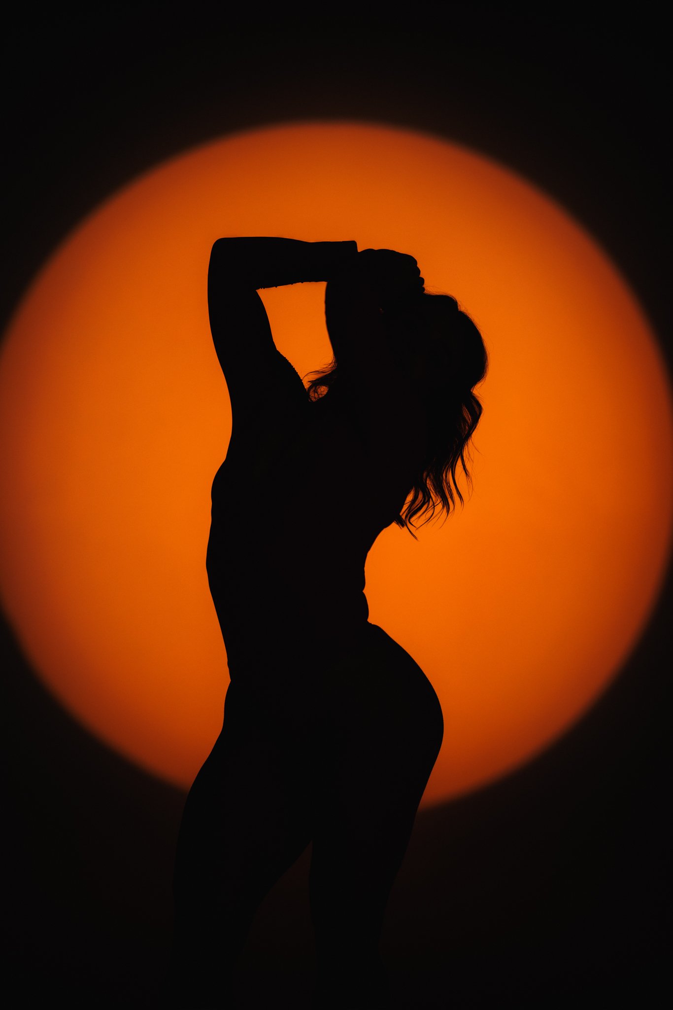 Silhouette of a woman with one hand behind her head, standing against an orange sunset or sunrise.