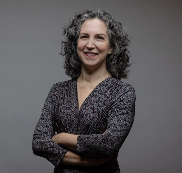Professional headshot of a woman with curly, shoulder-length silvery-black hair, smiling warmly at the camera with her arms crossed. She is wearing a dark, patterned long-sleeve top with a geometric design in muted red, blue, and cream tones. 