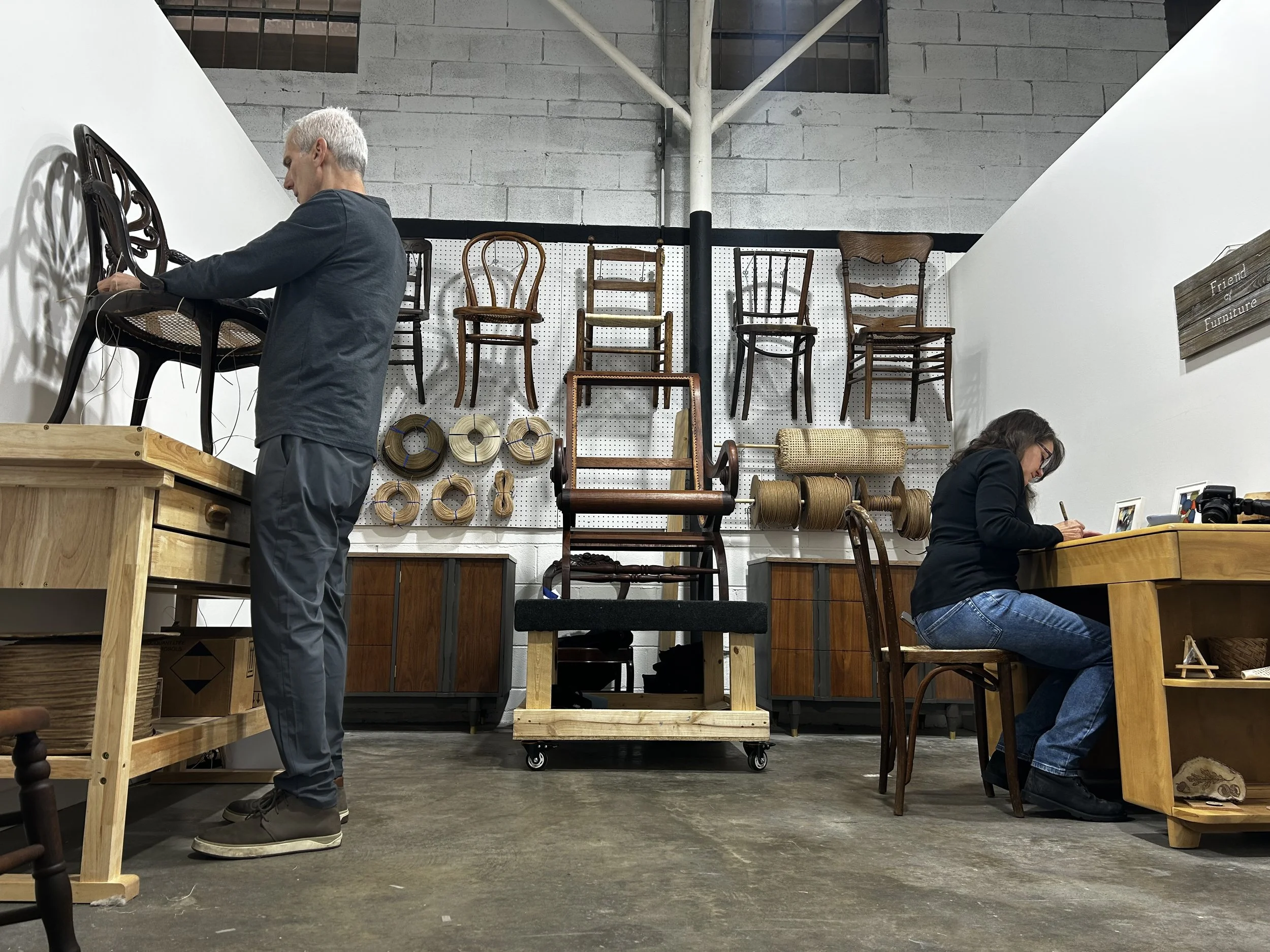 Furniture artisans repairing a wooden chair frame in Friend of Furniture's Asheville workshop, with chairs awaiting caning repair in background.