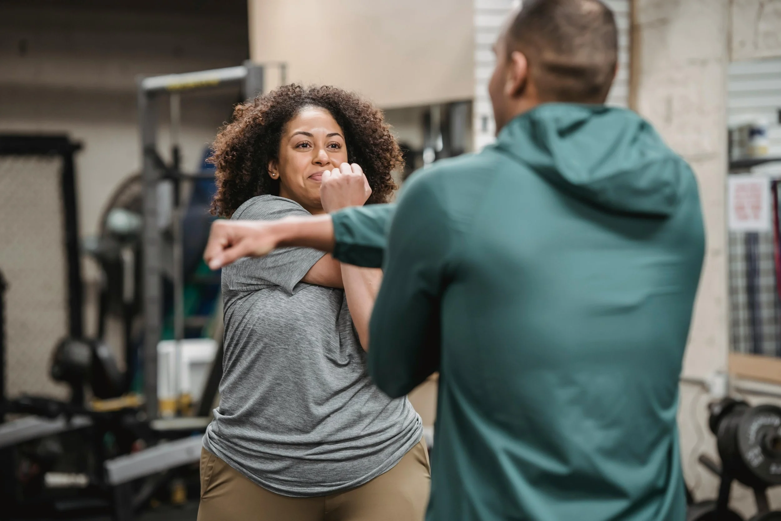 A woman with curly hair smiling and covering her mouth, sparring with a man at a gym.