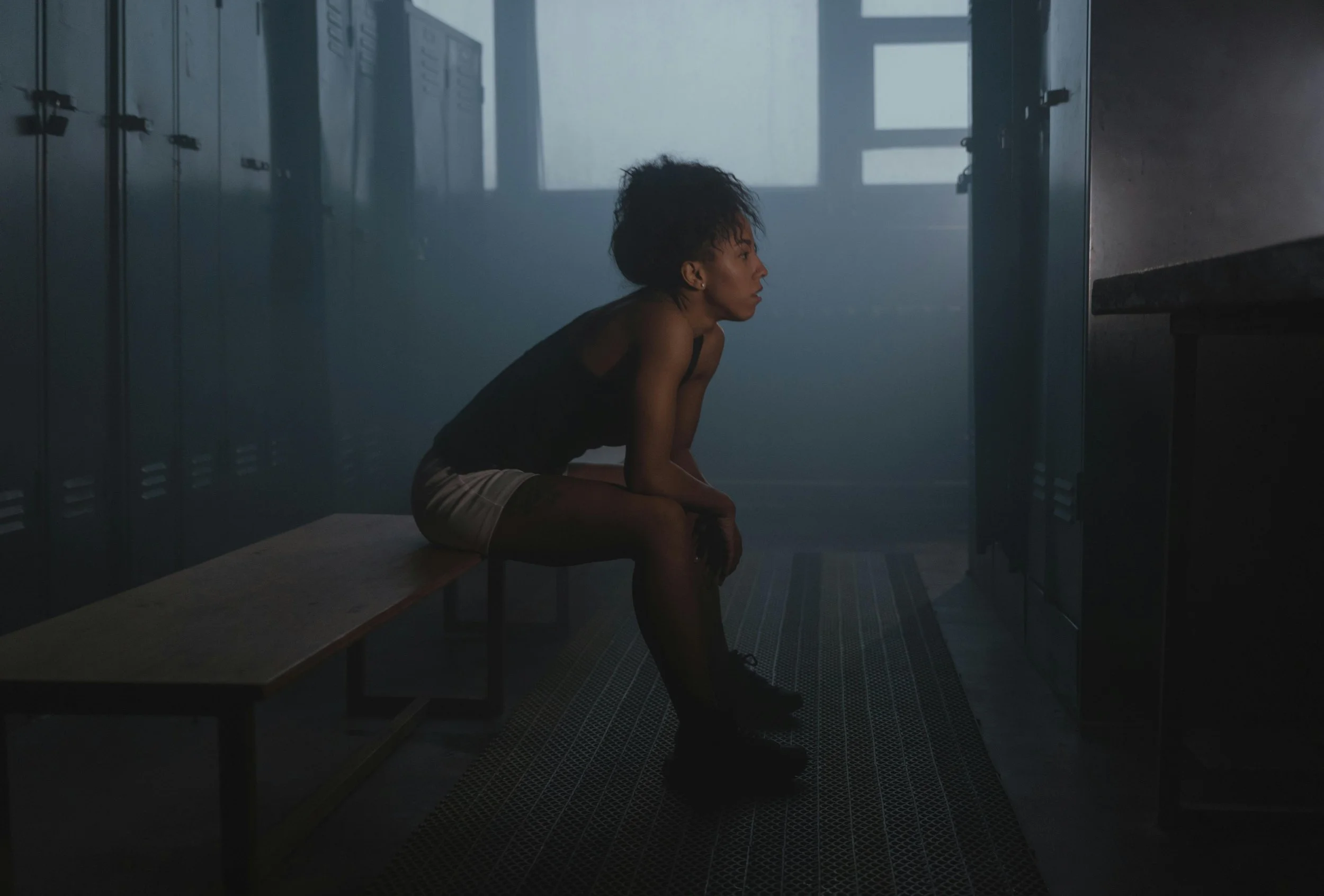 A young woman sitting on a wooden bench in a dimly lit locker room, dressed in a black tank top and beige shorts, with curly hair and black boots.
